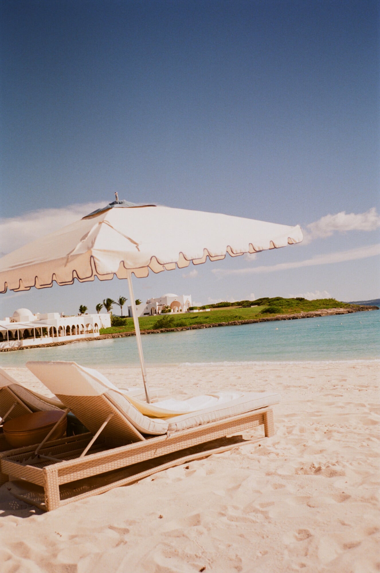 Cap Juluca, Anguilla — Cream umbrella and woven loungers on pristine sand beach with turquoise water and whitewashed coastal architecture.