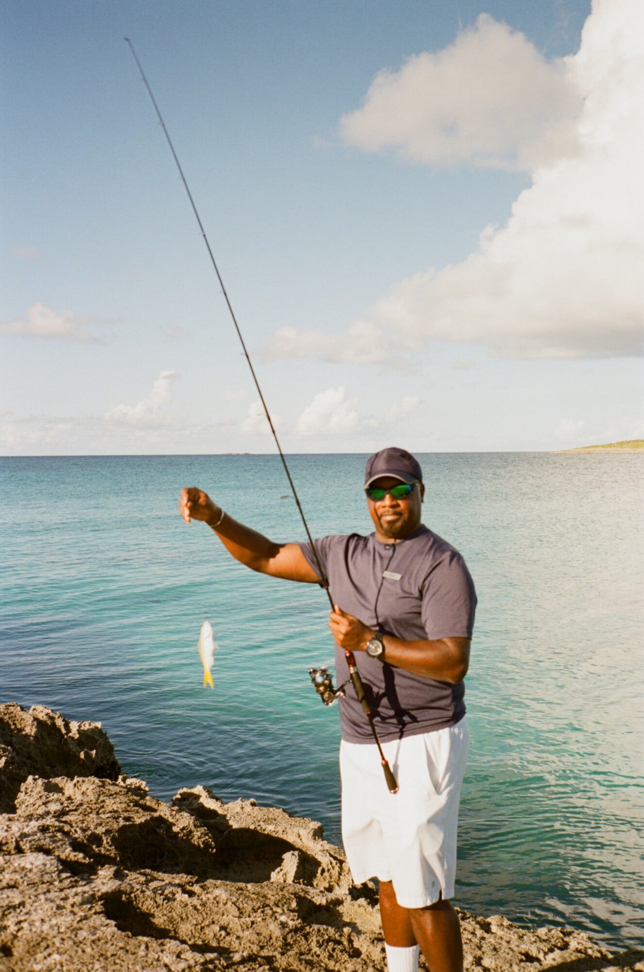 Fishing with Shawn — Man in sunglasses and cap holding fishing rod with catch in turquoise ocean waters off limestone rocky shore under blue sky.