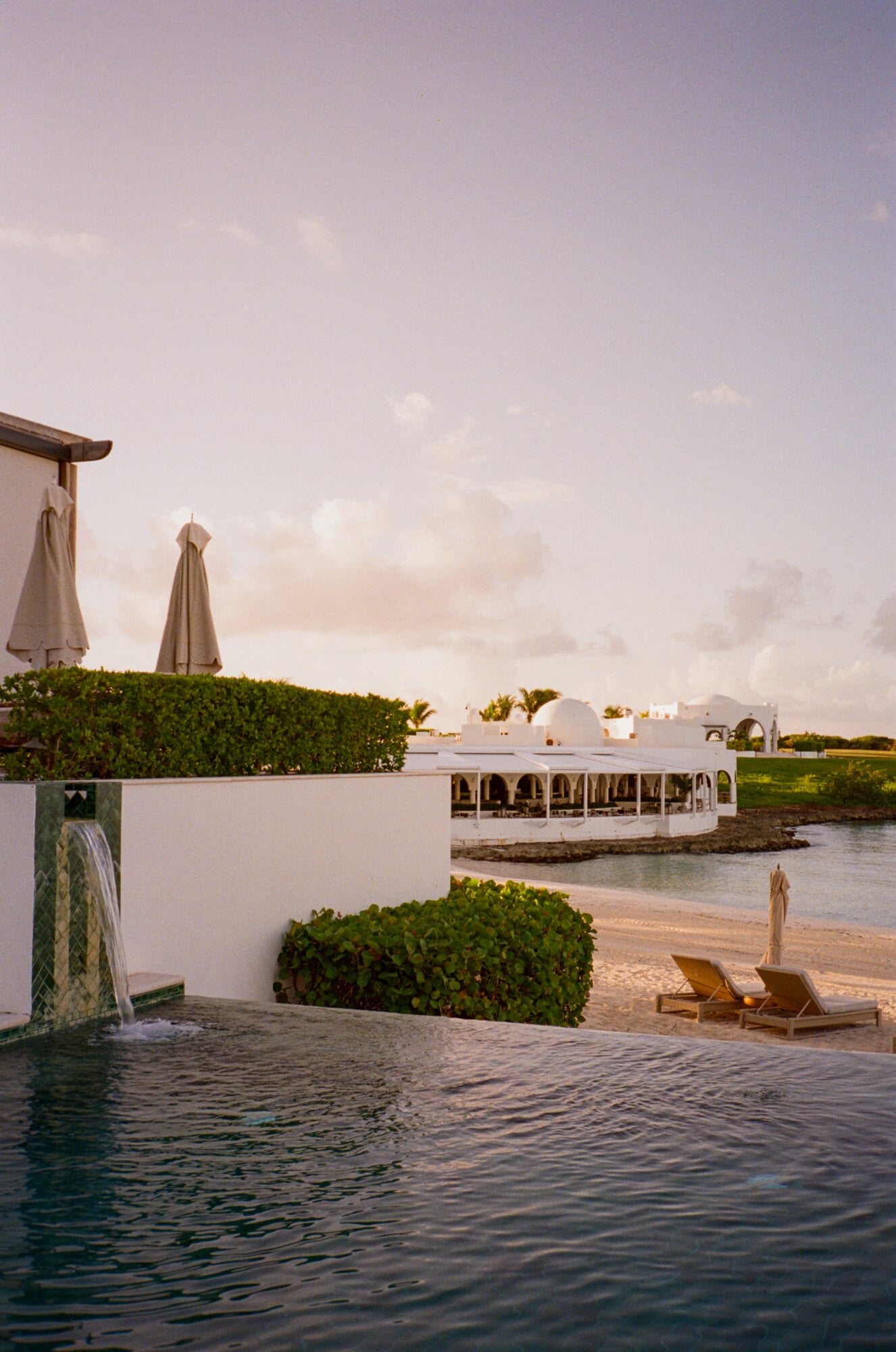 Cap Juluca, North America — Beachfront infinity pool overlooking lagoon with Moroccan-style pavilion, manicured hedges, lounge chairs, and whitewashed architecture at golden hour.