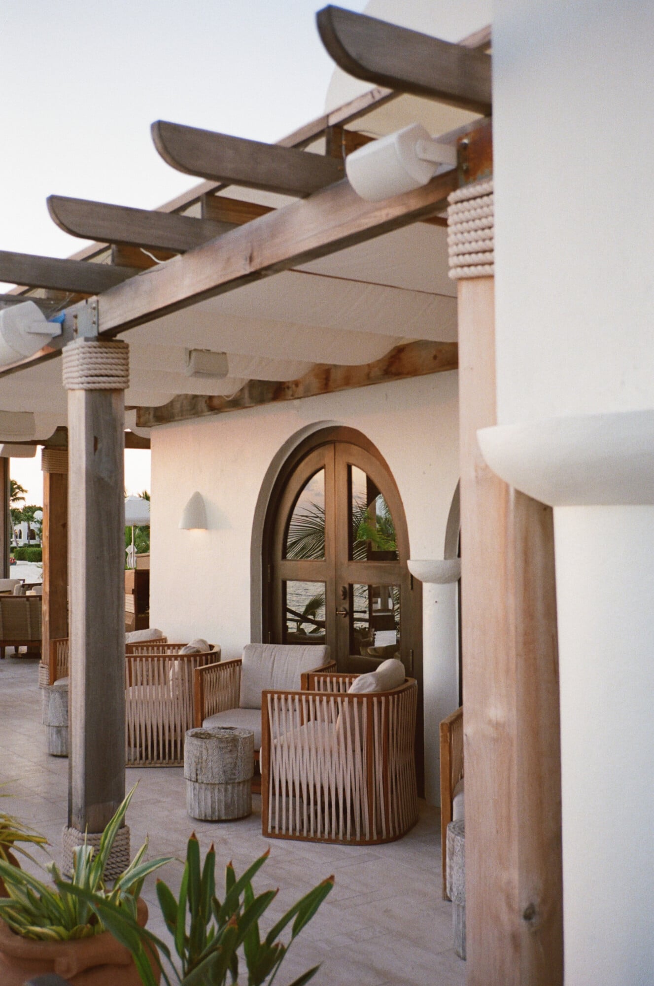 Cap Juluca, North America — Whitewashed courtyard with curved wooden archway, timber pergola, woven lounge chairs, and cone pendant lights at dusk.