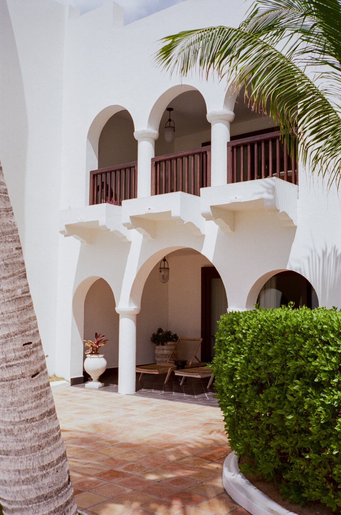 Cap Juluca, North America — Whitewashed arched colonnade with terracotta floor, burgundy railings, palm fronds, and manicured hedge in Mediterranean resort courtyard.