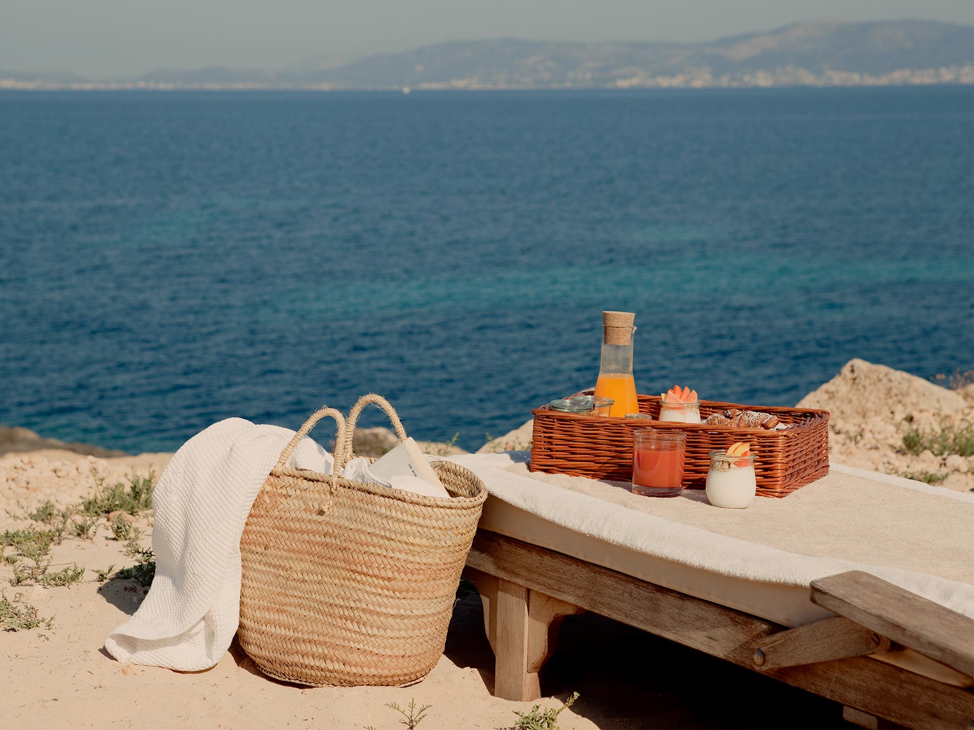 Cap Rocat, Balearic Islands — Woven basket with cream linen, wooden bench, picnic hamper and aperitif overlooking Mediterranean coast at golden hour.