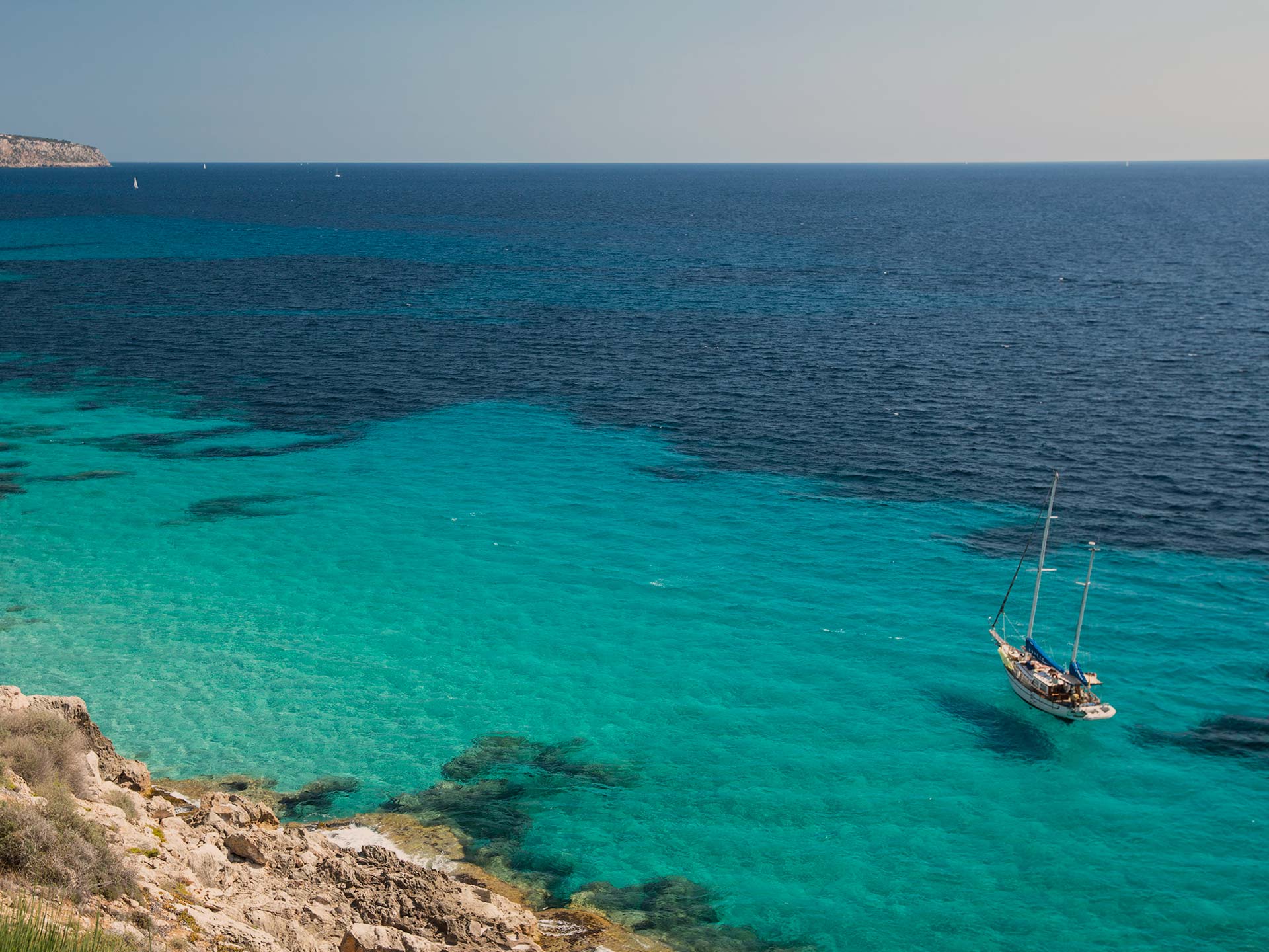 Cap Rocat, Balearic Islands — Sailboat anchored in crystalline turquoise waters off rocky coastline under clear Mediterranean sky.