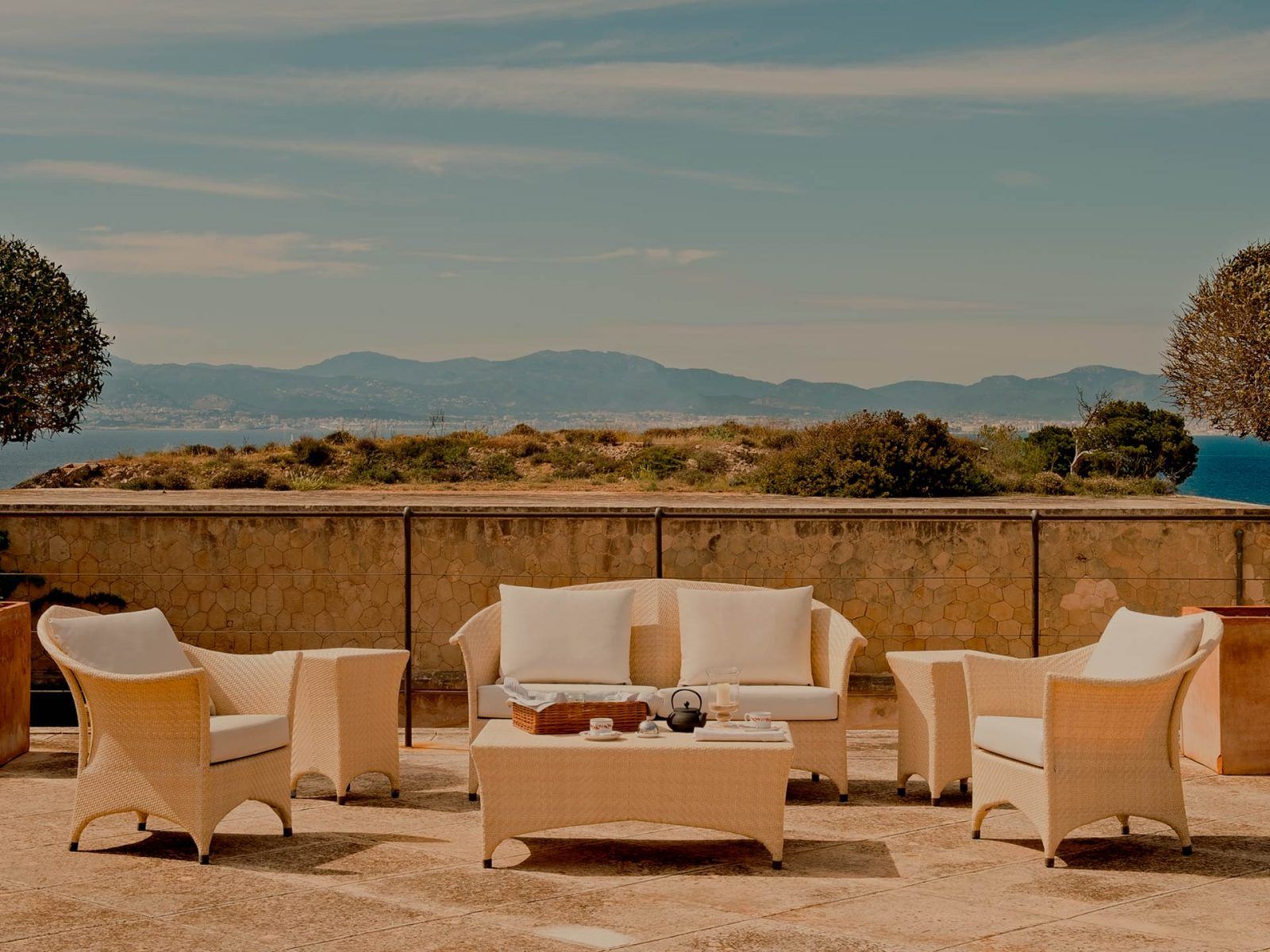 Cap Rocat, Balearic Islands — Woven lounge chairs with cream cushions arranged on stone terrace overlooking Mediterranean Sea and distant mountains.