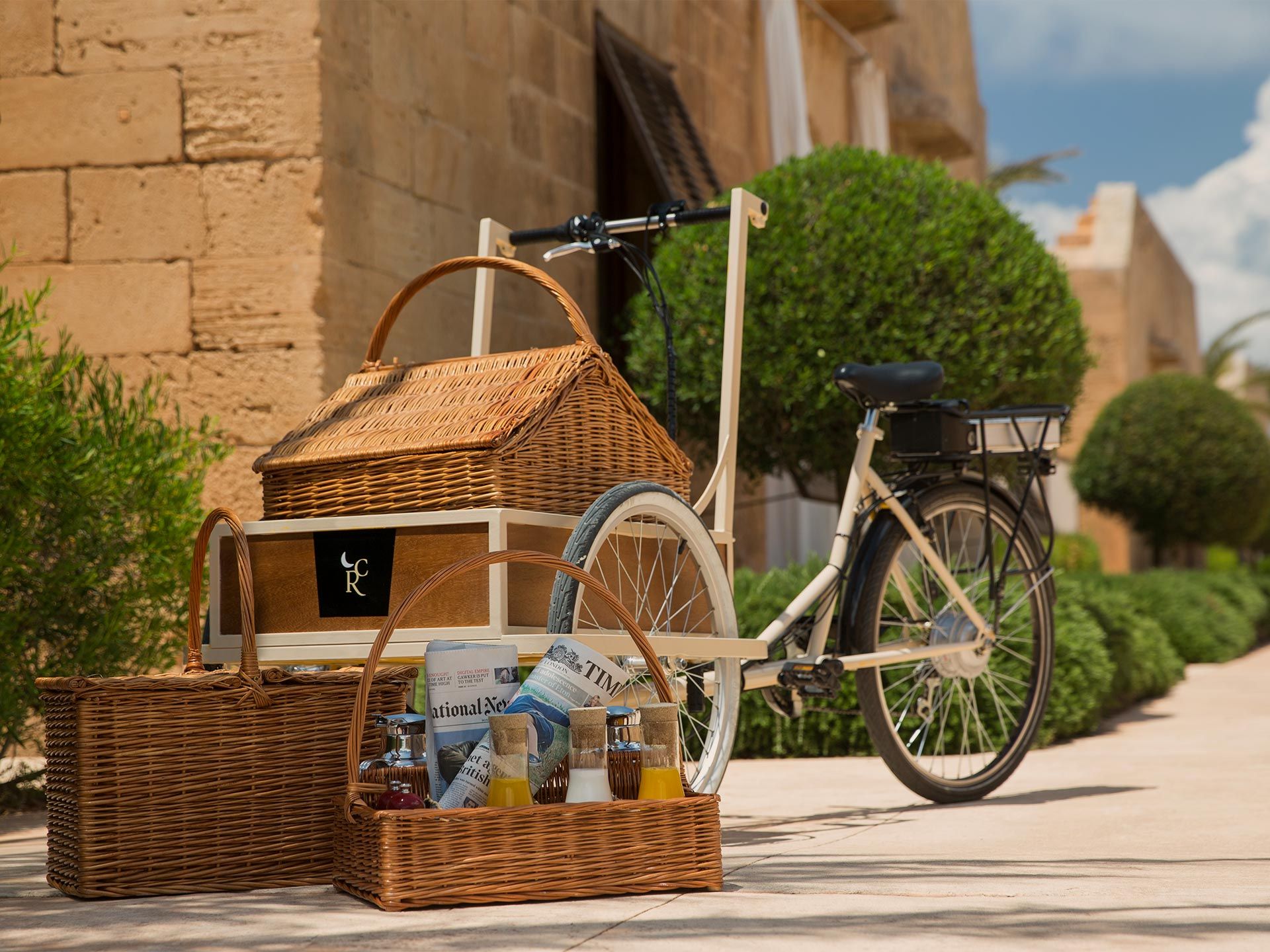 Cap Rocat, Balearic Islands — Wicker picnic baskets and cream bicycle with cargo carrier arranged on stone terrace against Mediterranean stone fortress architecture.