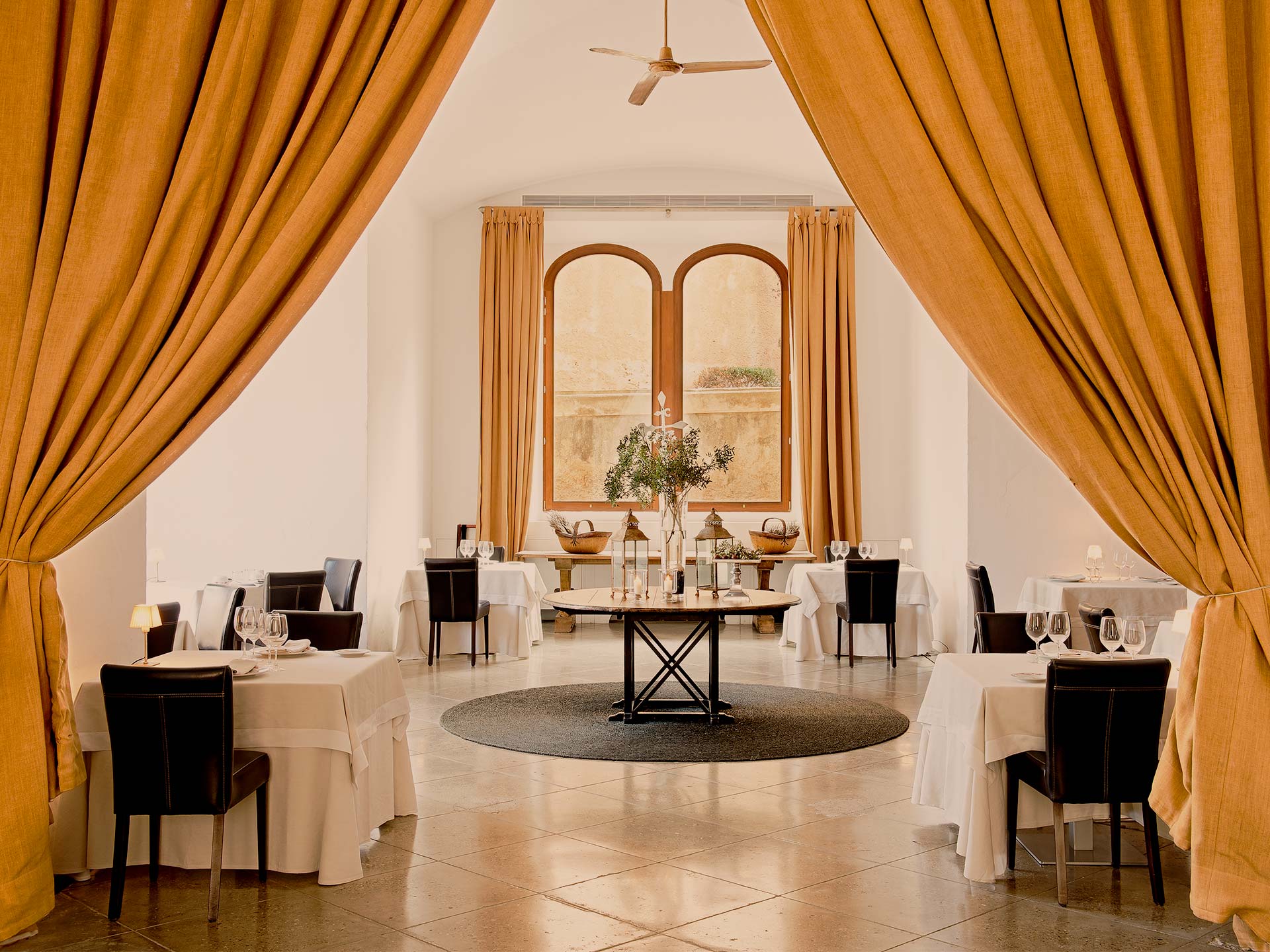 La Fortaleza, Balearic Islands, Spain — Ochre curtains frame arched windows overlooking stone terraces in minimalist dining room with cream linens and black chairs.
