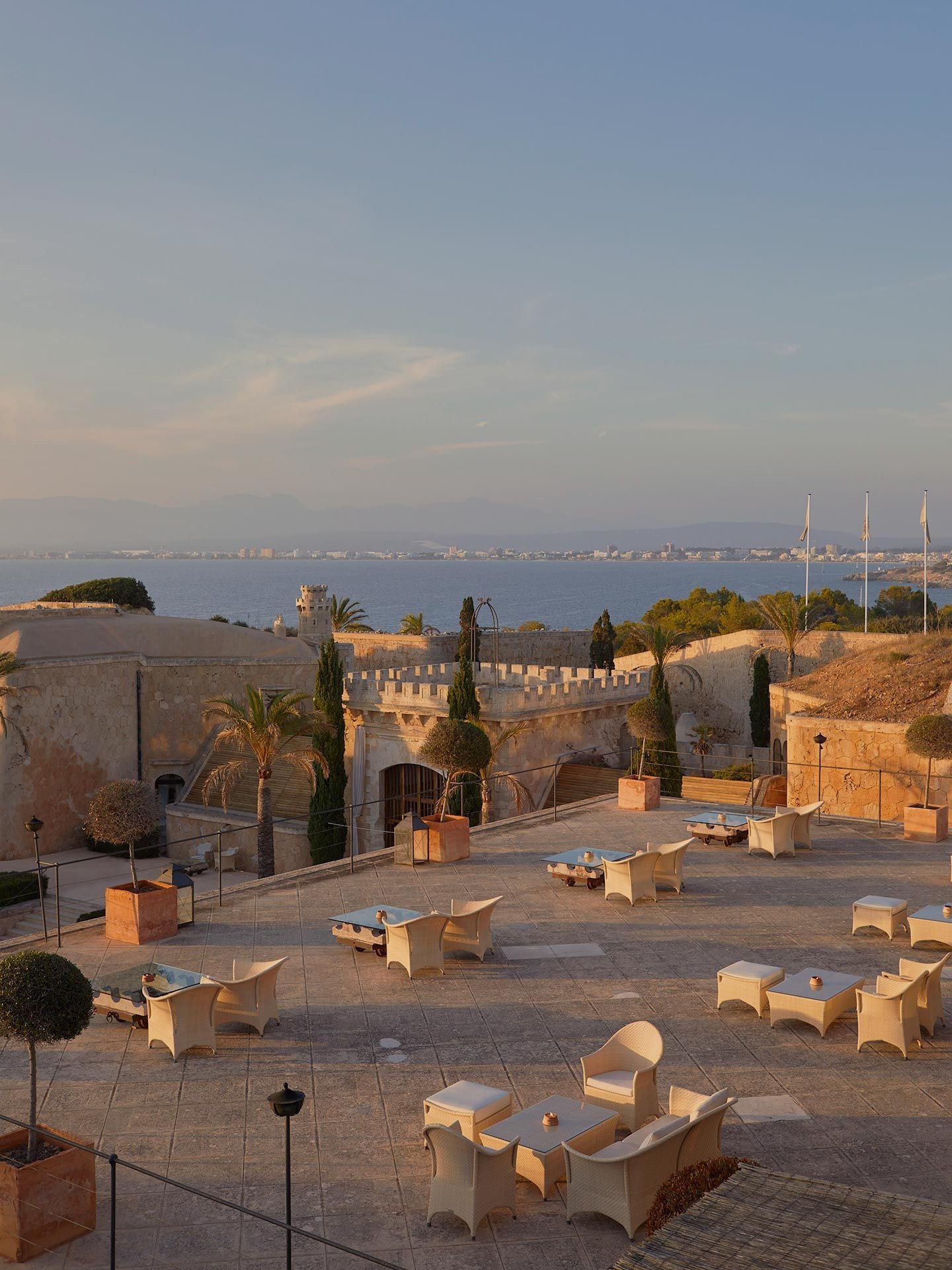 Cap Rocat, Balearic Islands, Spain — Golden-hour courtyard with cream lounge furniture, historic stone fortress architecture, Mediterranean sea and coastline beyond.