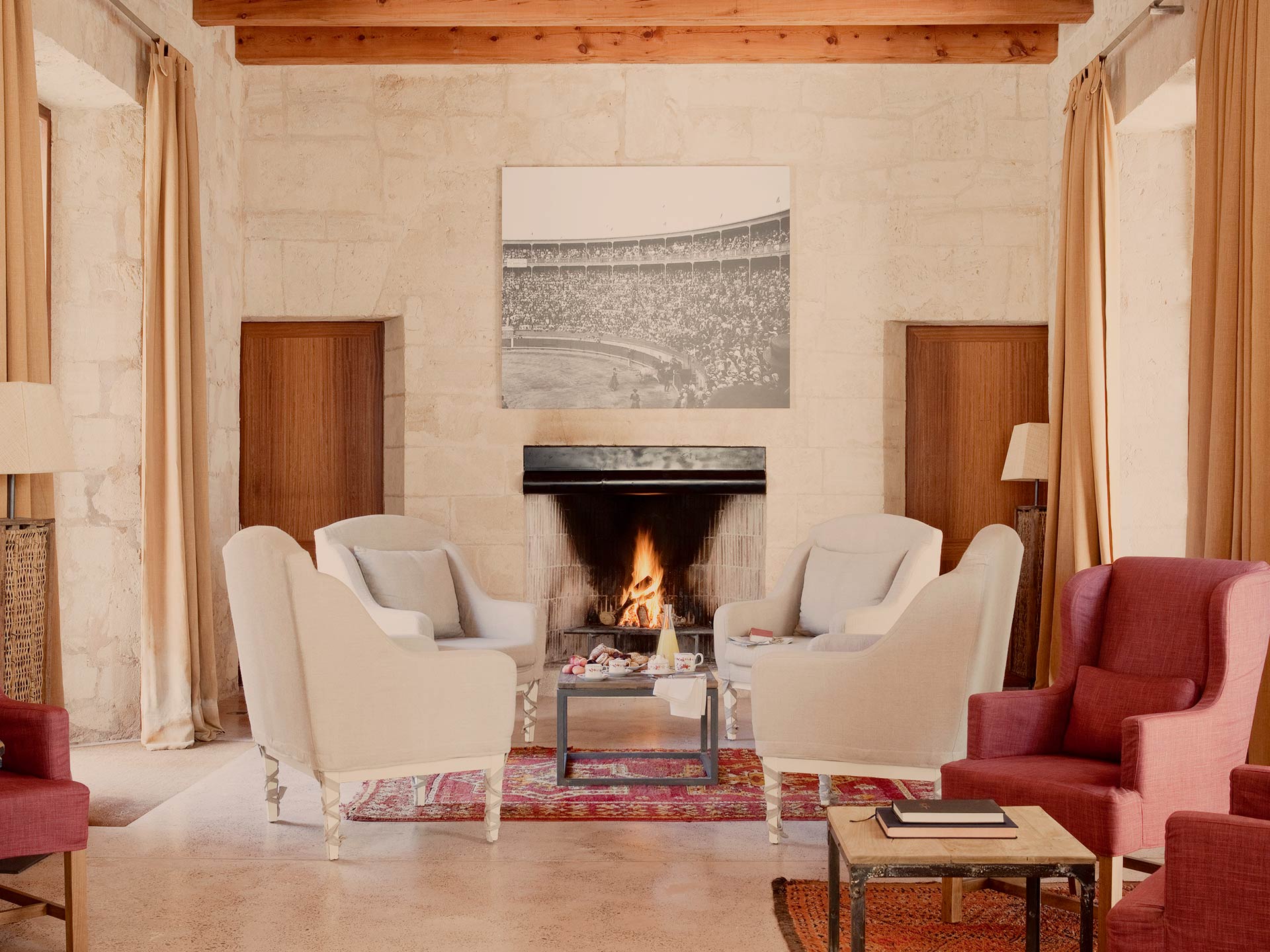 Cap Rocat, Balearic Islands — Limestone fireplace lounge with cream armchairs, red wingback seating, wooden beams, and vintage stadium photograph.