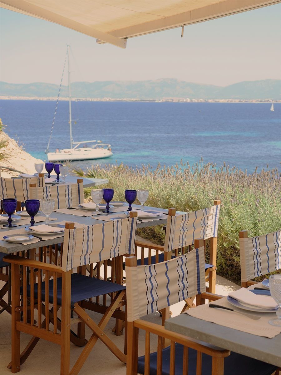 Sea Club, Balearic Islands — Seaside dining terrace with striped linen chairs, wooden table, blue glassware, sailboat anchorage, and Mediterranean coastline under canvas shade.