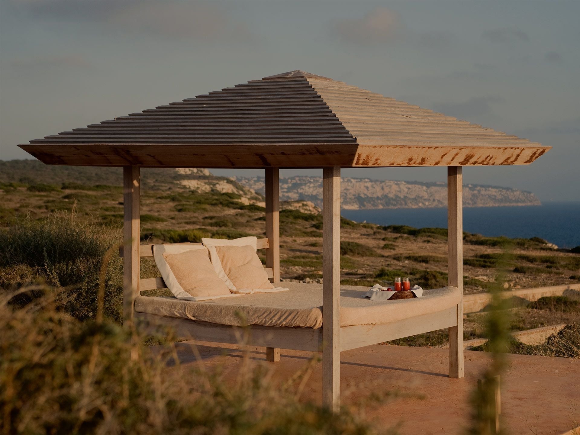 Cap Rocat, Balearic Islands — Wooden pergola bed pavilion with cream cushions overlooking Mediterranean coastline at sunset.