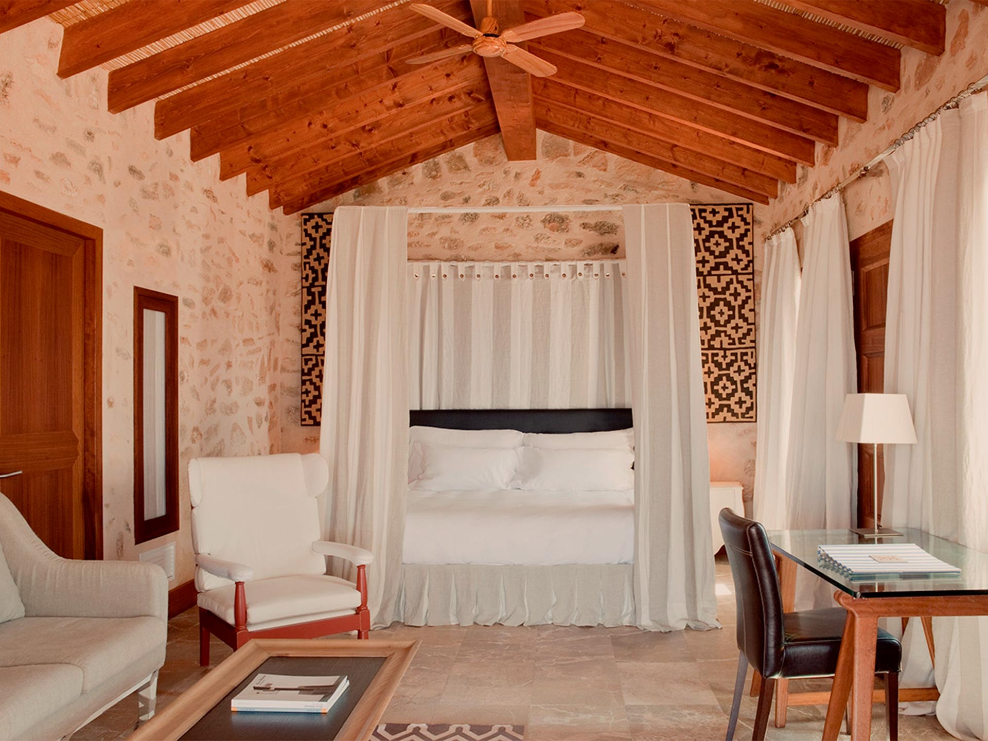 Cap Rocat, Balearic Islands — Canopied bed with cream curtains in stone bedroom with exposed wood beams and geometric textile panels.