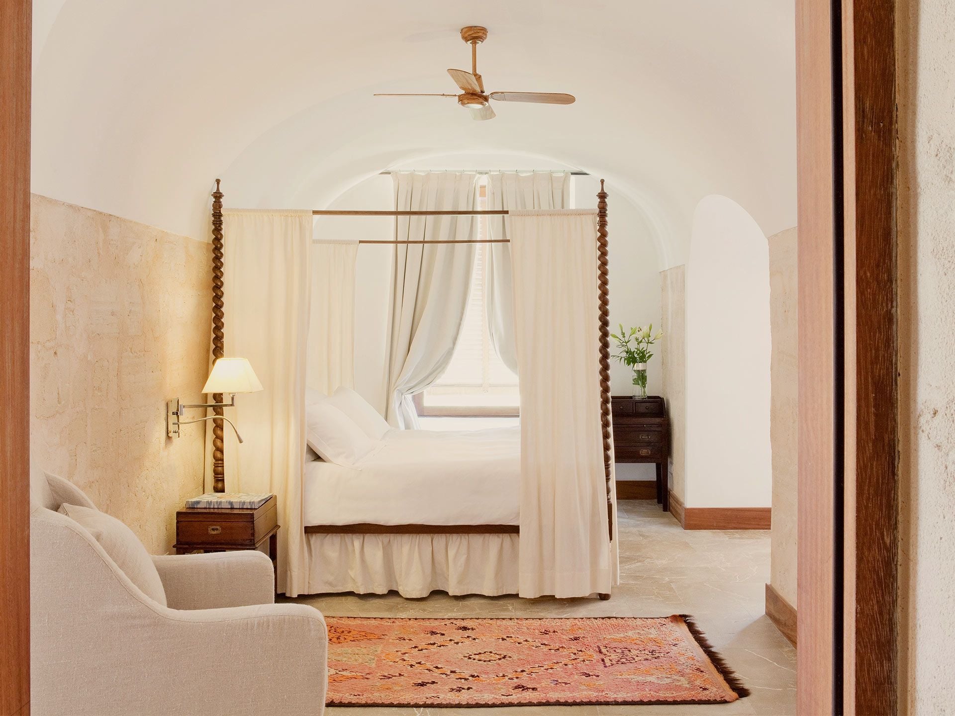 Cap Rocat, Balearic Islands — Canopy bed with cream linen drapes, arched stone walls, wooden ceiling fan, terracotta tile floor, minimalist Mediterranean luxury bedroom.