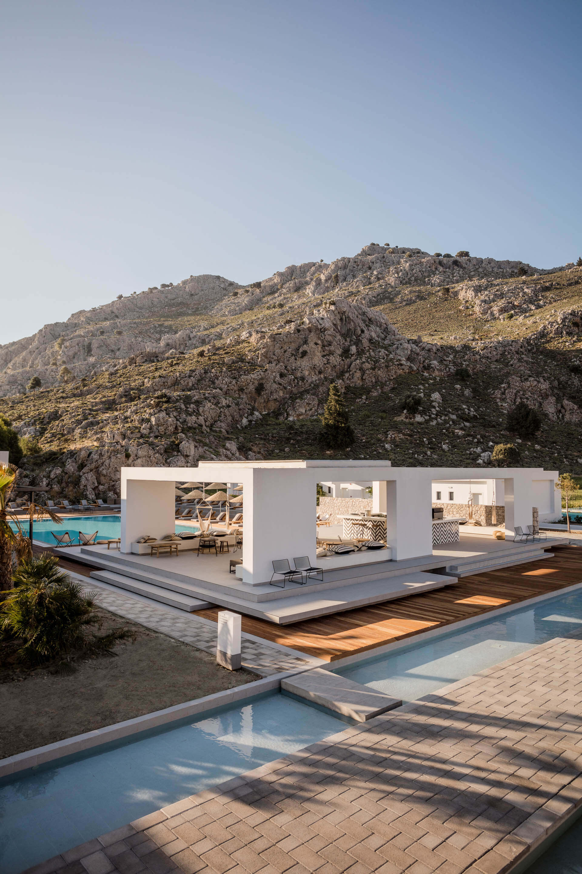 Casa Cook Rhodes, Greece — minimalist white pavilion with geometric cutouts overlooking turquoise pool, rocky hillside, and Mediterranean landscape in golden afternoon light.