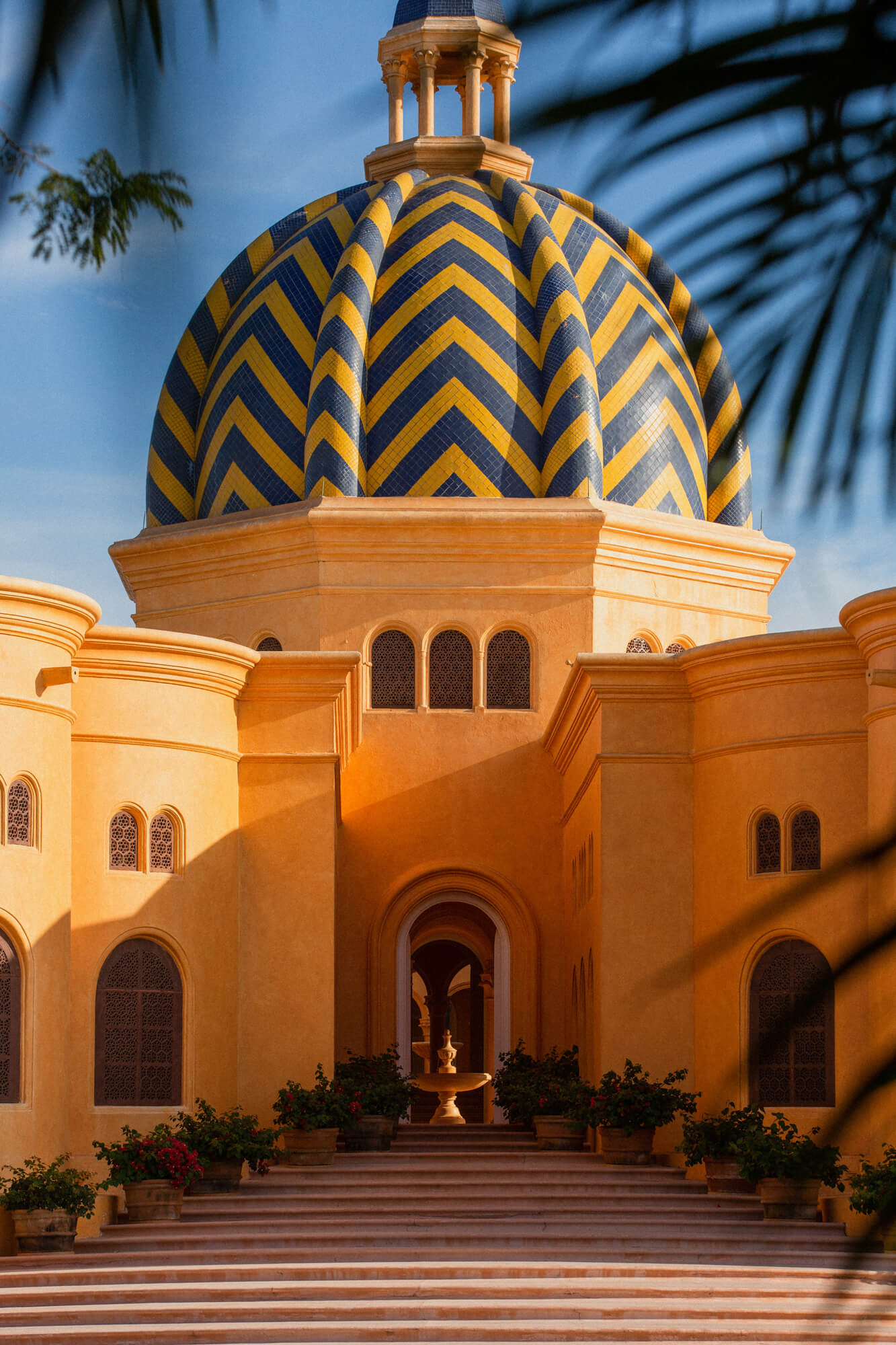 Cuixmala, Mexico — Ochre-washed chapel with blue and gold chevron-patterned dome, arched windows, and palm-shaded courtyard in brilliant daylight.