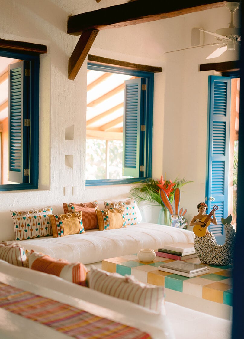 Casa Gomez, Mexico — Sunlit living room with teal louvered doors, wooden beams, white walls, striped pillows, and colorful geometric floor tiles.