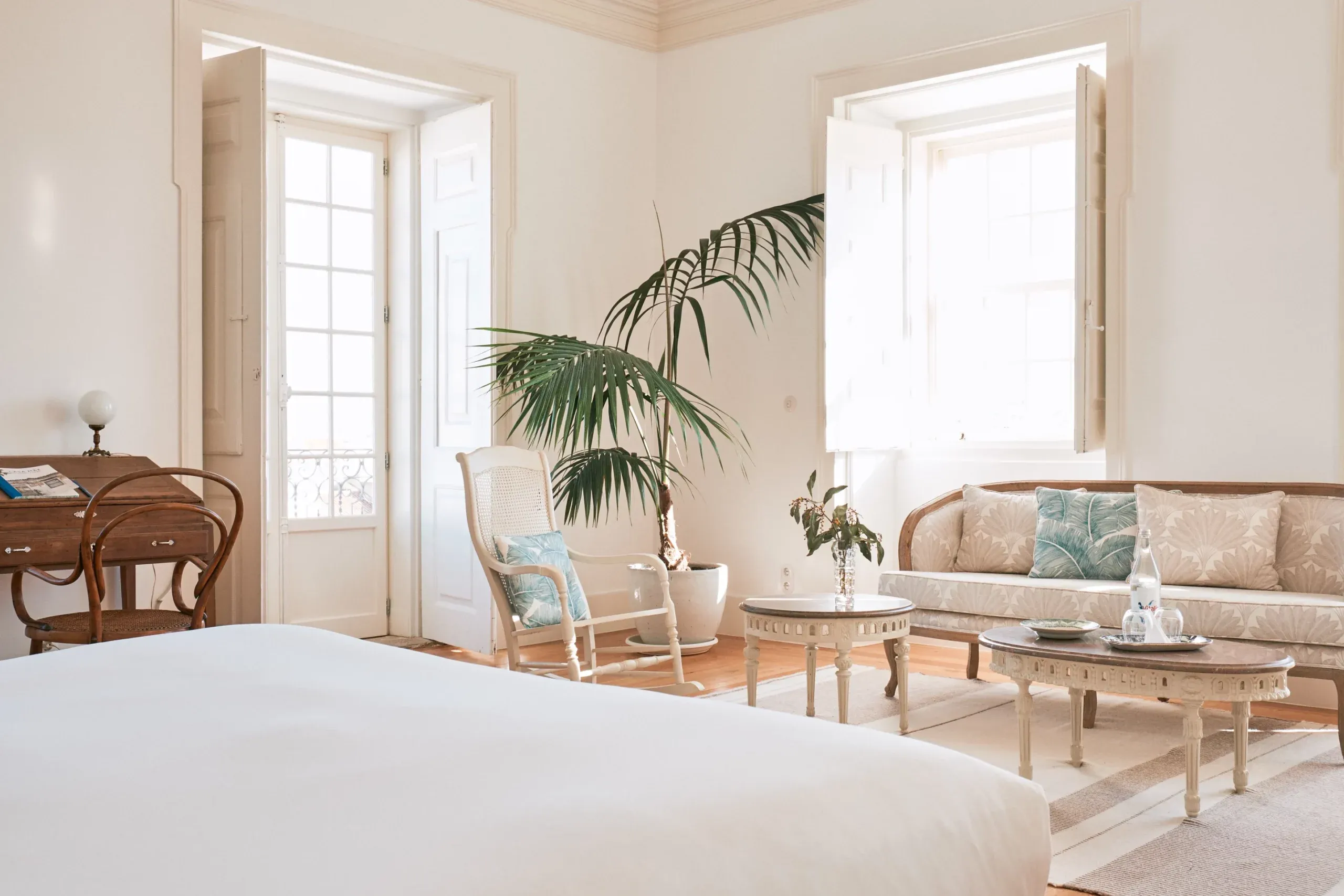 Casa Mãe, Europe — Bright bedroom suite with cream furnishings, palm plant, light wood furniture, and French doors opening to dual lounge areas with botanical accents.