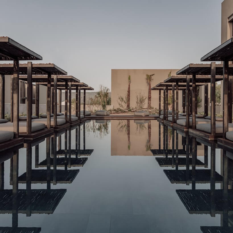 Casa Cook Samos, Greece — Minimalist poolside pavilions with dark pergolas reflected in still water, palm trees and beige architecture under overcast sky.