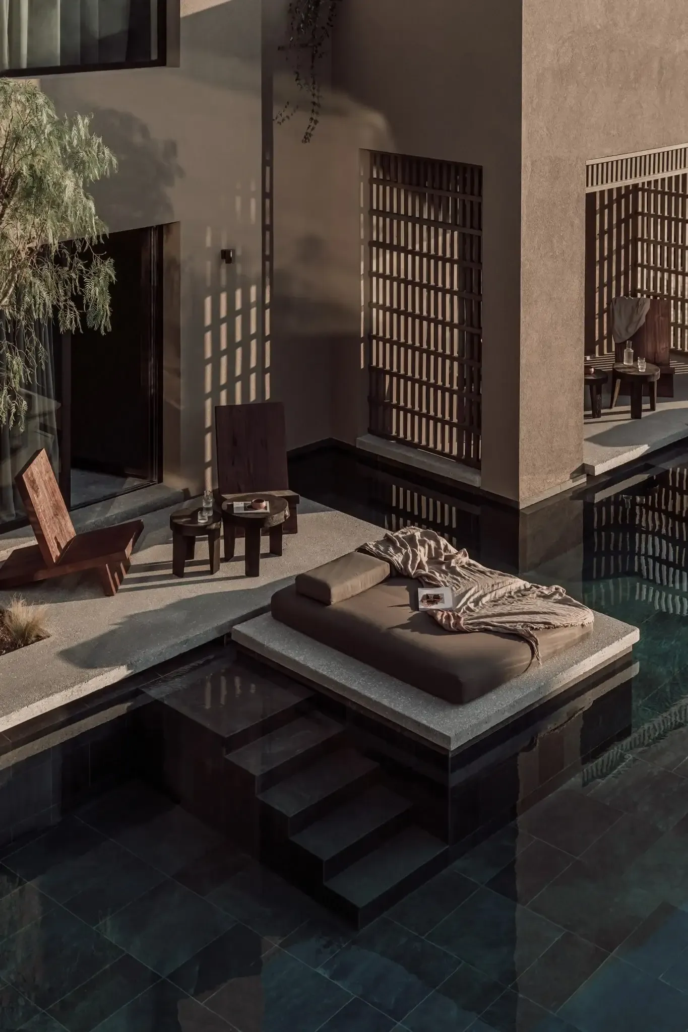 Casa Cook Samos, Greece — Minimalist courtyard with dark wooden lattice screens, reflecting pool, stone platform with cushioned daybed, and dappled sunlight casting geometric shadows on warm terracotta walls.
