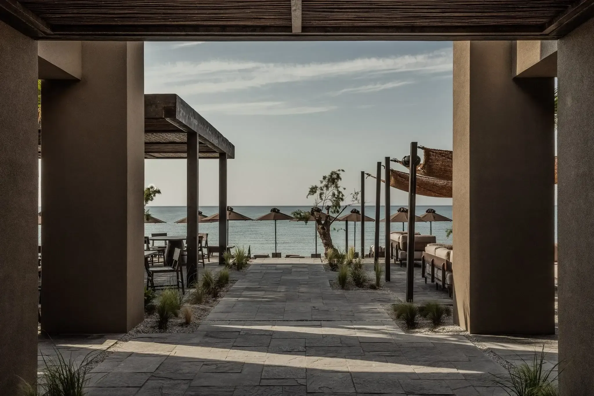 Casa Cook Samos, Greece — Covered terrace with dark wooden pergolas framing turquoise Aegean Sea view, stone paving, minimalist coastal architecture.