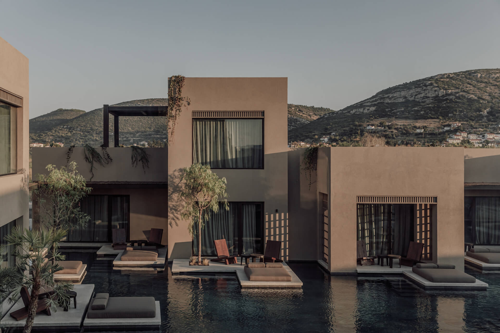 Casa Cook Samos, Greece — Modernist resort courtyard with terracotta facades, reflecting pools, dark timber screens, and arid hillside backdrop at golden hour.