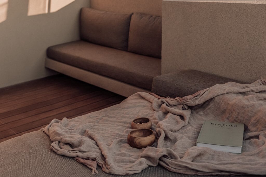 Casa Cook Samos, Greece — Minimalist bedroom with linen bedding, wooden bowls, and book on bed facing brown upholstered sofa in warm natural light.