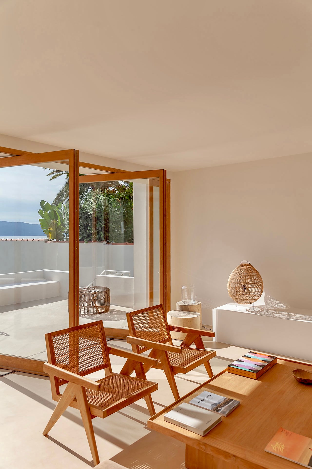 Casa Santa Teresa, Europe — Minimalist terrace with wooden mid-century chairs, woven pendant lamp, open doors framing Mediterranean views, cream walls and natural light.
