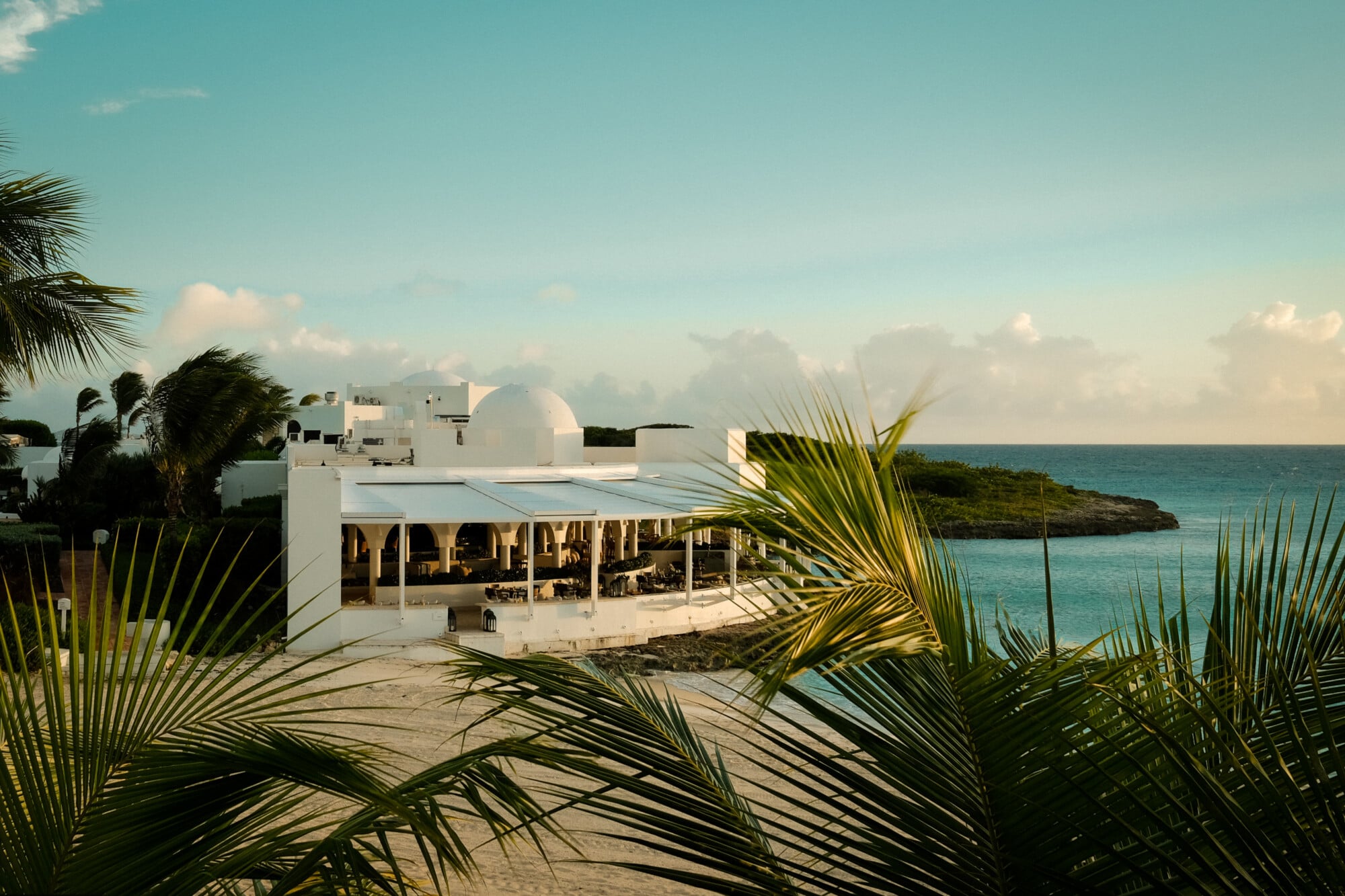 Cip's by Cipriani, North America — Modernist white pavilion with open veranda overlooking turquoise ocean, palm fronds in foreground, coastal architecture at golden hour.