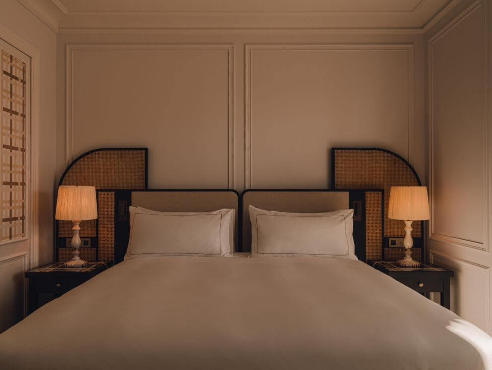 Carlton Cannes, Cannes, France — Neutral-toned bedroom with symmetrical black-framed rattan headboards, cream pillows, brass table lamps, and paneled walls in luxury hotel design.