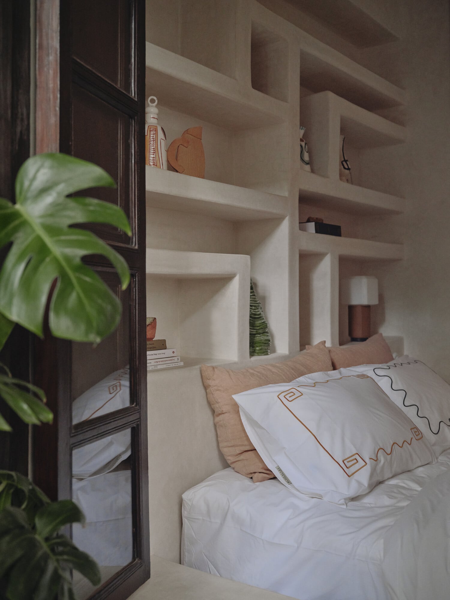 Rosemary, Africa — Minimalist bedroom with built-in shelving, white bedding with geometric trim, dark wood door, and monstera plant foreground.