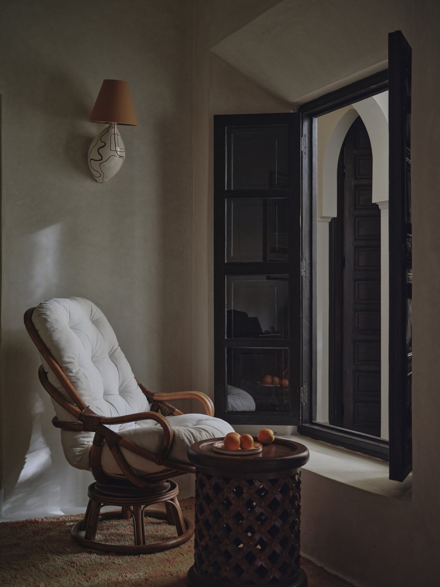 Rosemary, Africa — Rattan lounge chair with white cushions beside carved wooden side table, framed by black-trimmed arched doorway and minimalist wall sconce, warm Mediterranean light.
