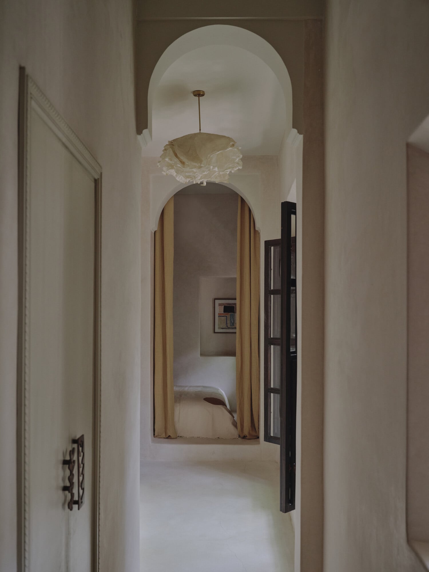 Rosemary, Africa — Arched hallway with pendant light, cream walls, dark wooden door, and mirror reflecting bed through doorway.