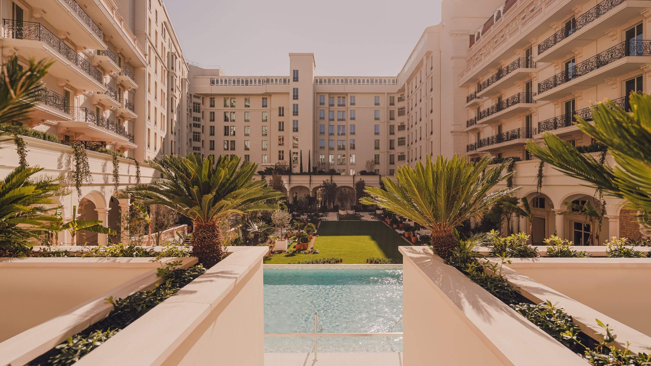 Carlton Cannes, Cannes, France — Neoclassical courtyard with reflecting pool, palm trees, manicured lawn, and cream-colored belle époque architecture.