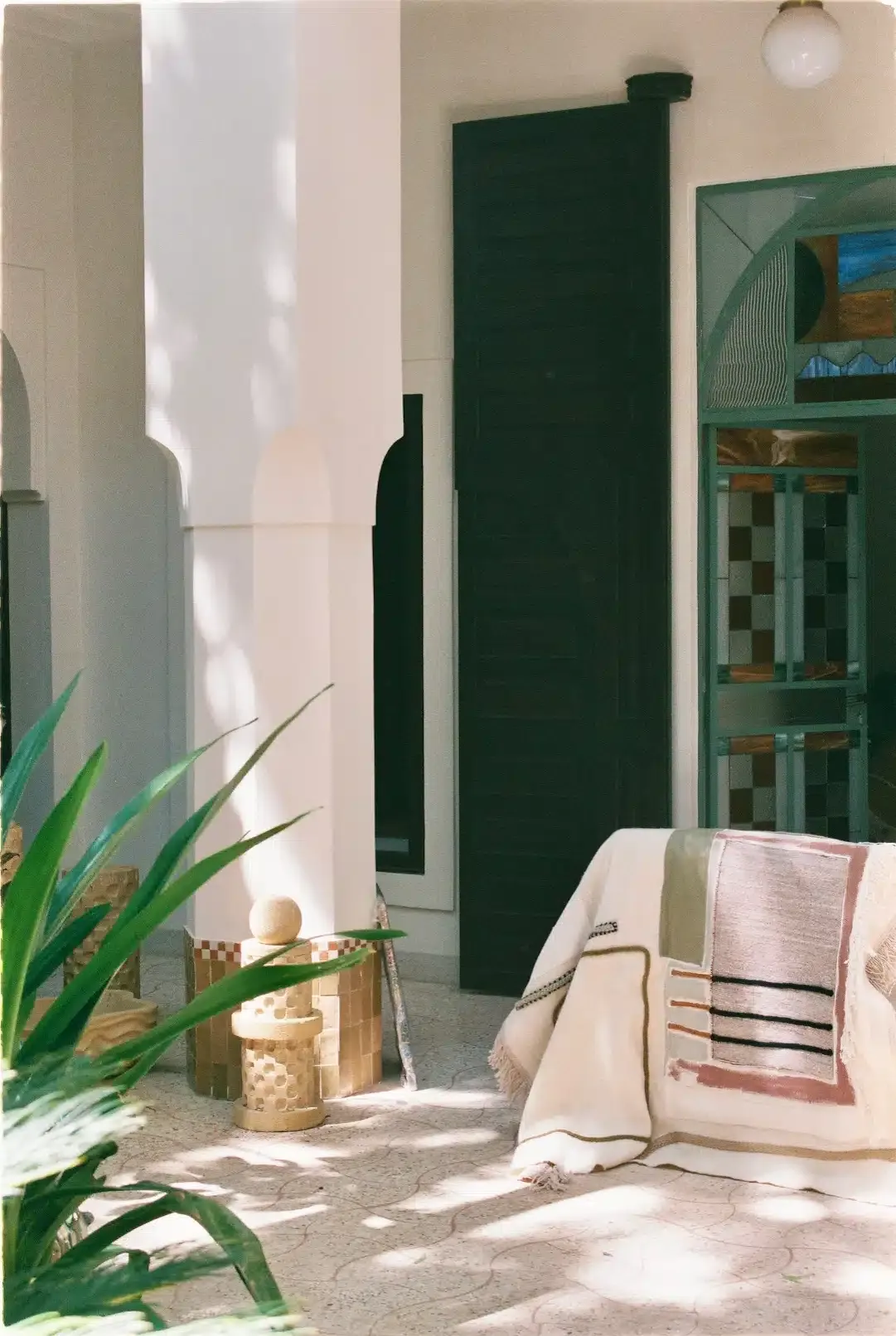 Rosemary, Africa — Minimalist courtyard with dark green door, whitewashed walls, natural light, woven baskets, green plants, and folded textiles on bench.