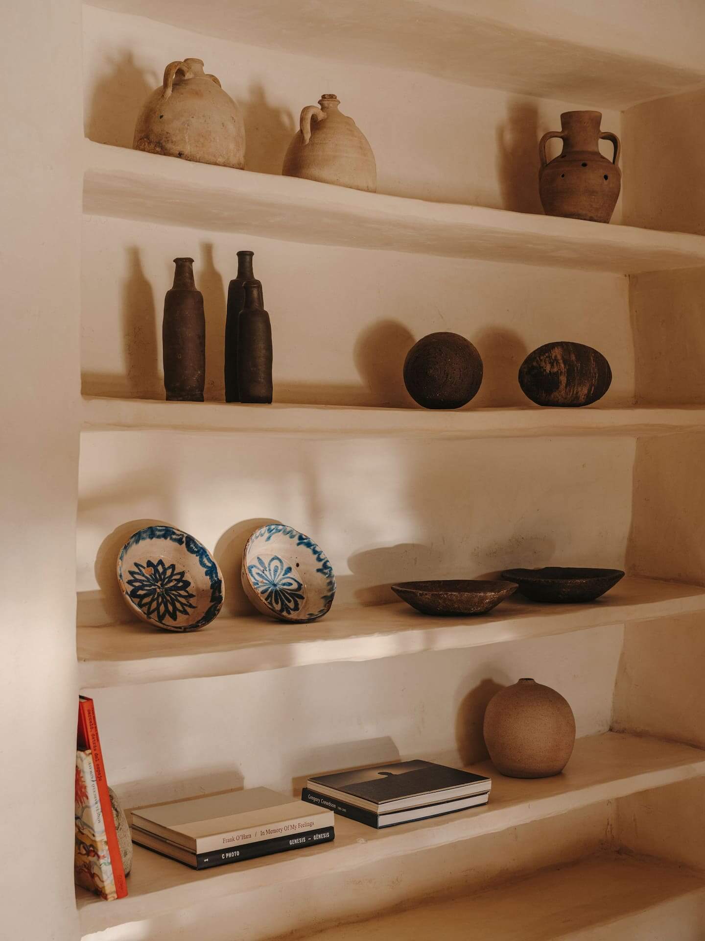 Can Lluïssó, Balearic Islands, Spain — Recessed wooden shelving displaying terracotta vessels, dark ceramics, blue-patterned bowls, and books in warm natural light.