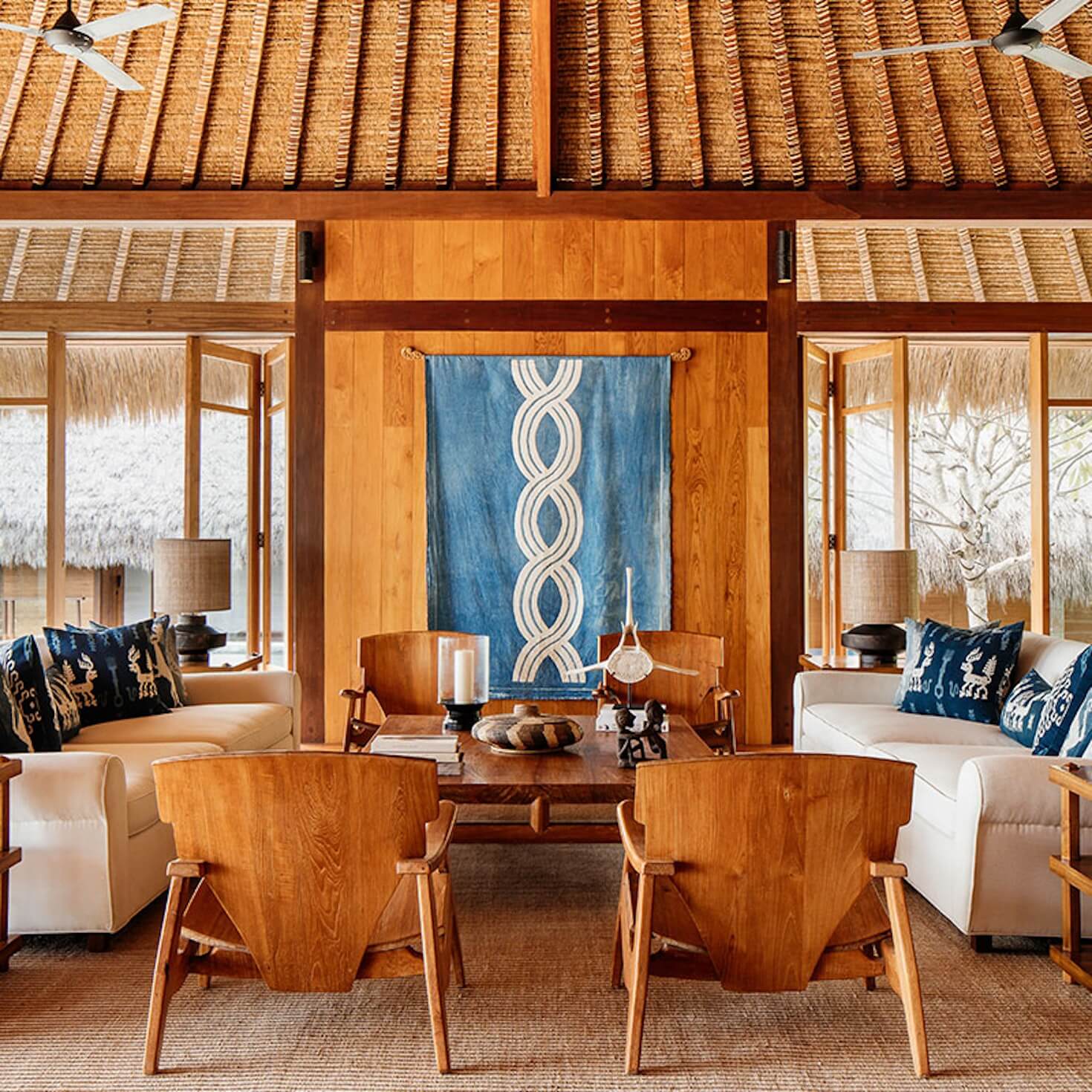 NIHI Sumba, Asia — Thatched pavilion living room with teak wood furnishings, blue textile art, cream upholstered seating, and ocean views through floor-to-ceiling windows.