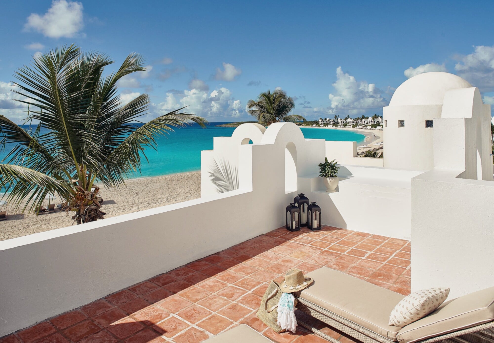 Cap Juluca, North America — Whitewashed terrace with curved architecture overlooking turquoise Caribbean beach, terra-cotta tiles, palm fronds, lounge seating, coastal luxury resort design.