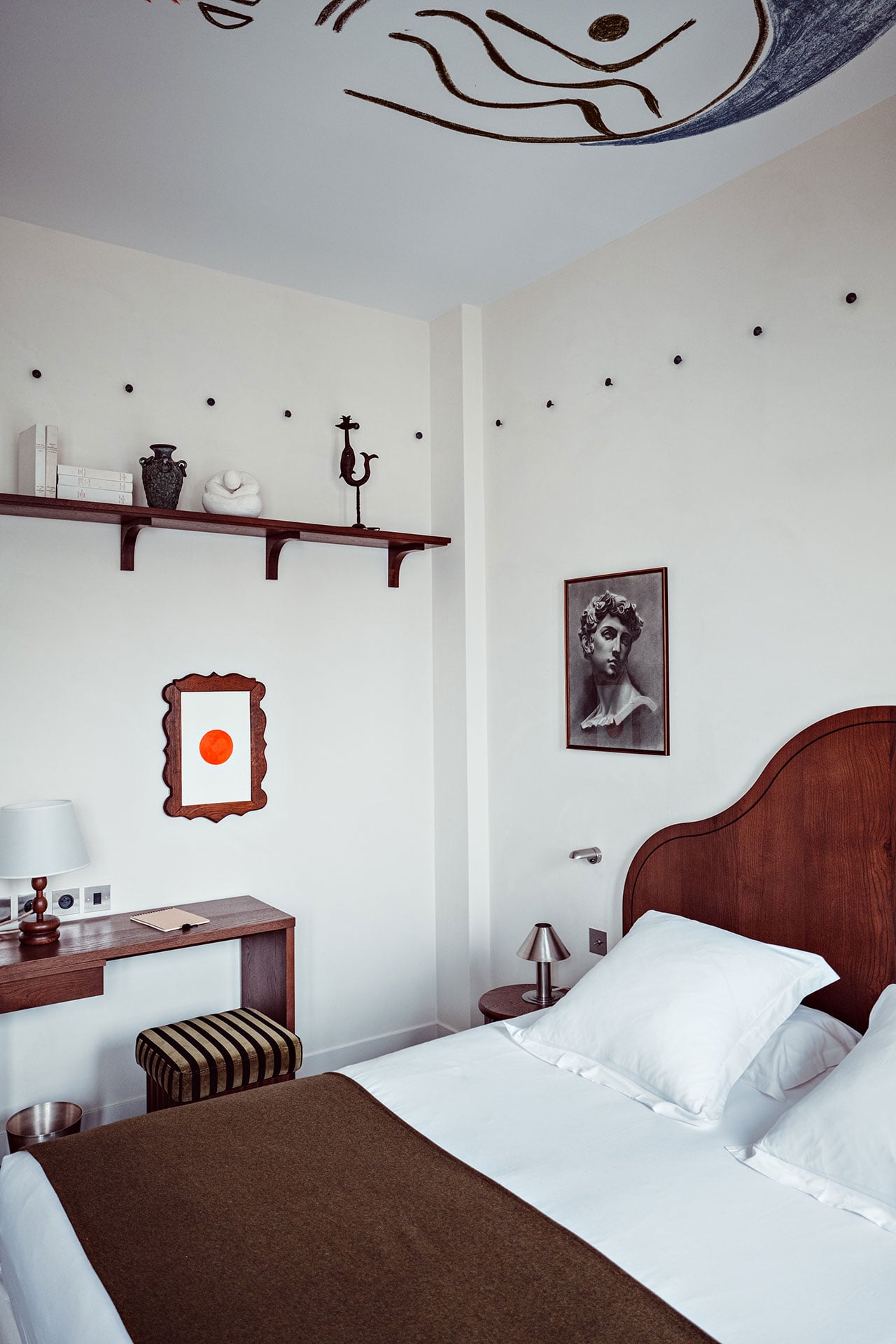 Académies et des Arts, Europe — Minimalist bedroom with burgundy wooden bed frame, white linens, dark wood desk, framed classical portrait, floating shelf, and abstract ceiling mural.