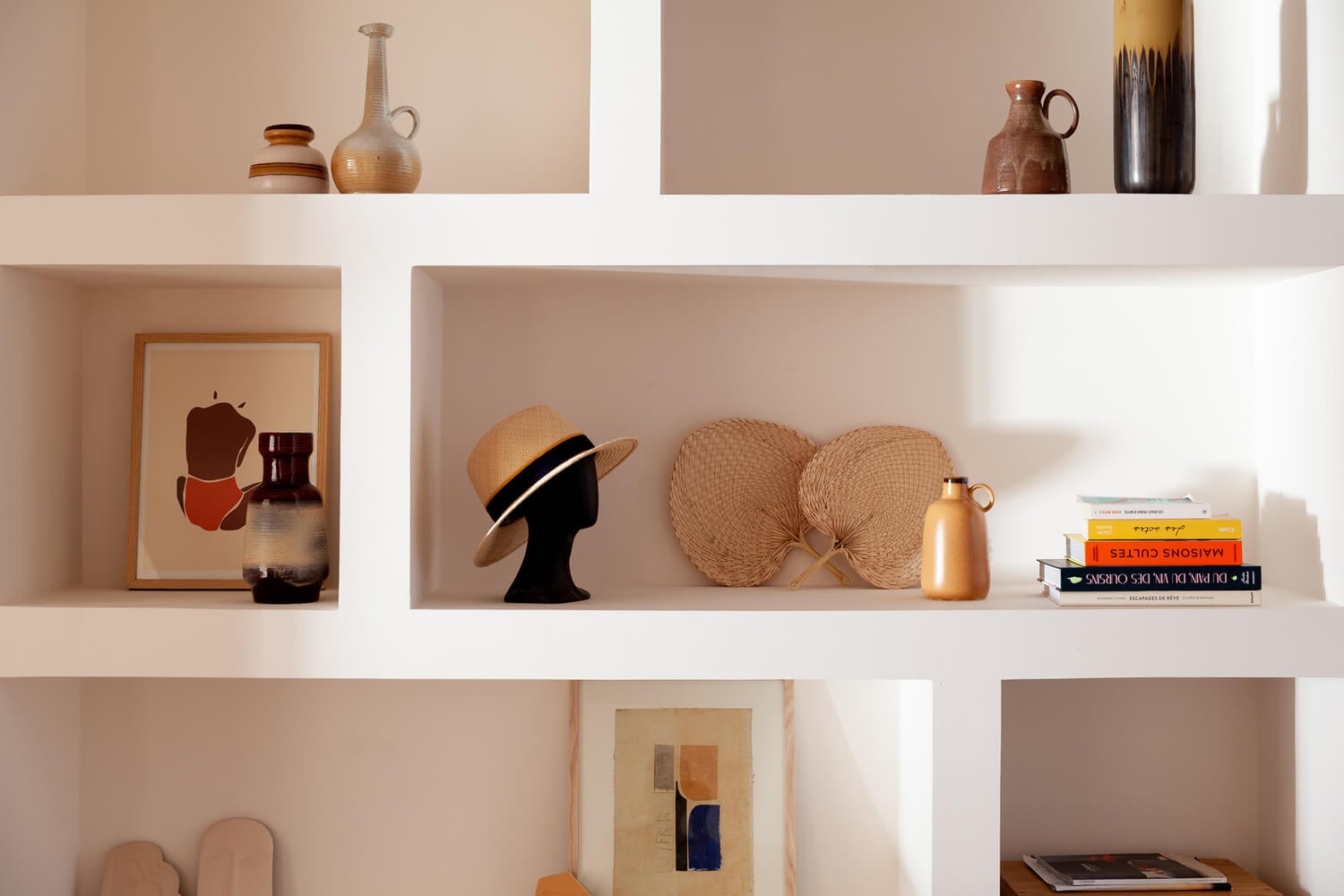 Casa Santa Teresa, Europe — White shelving with curated ceramics, straw hats, brass vessels, artwork and books in warm natural light.