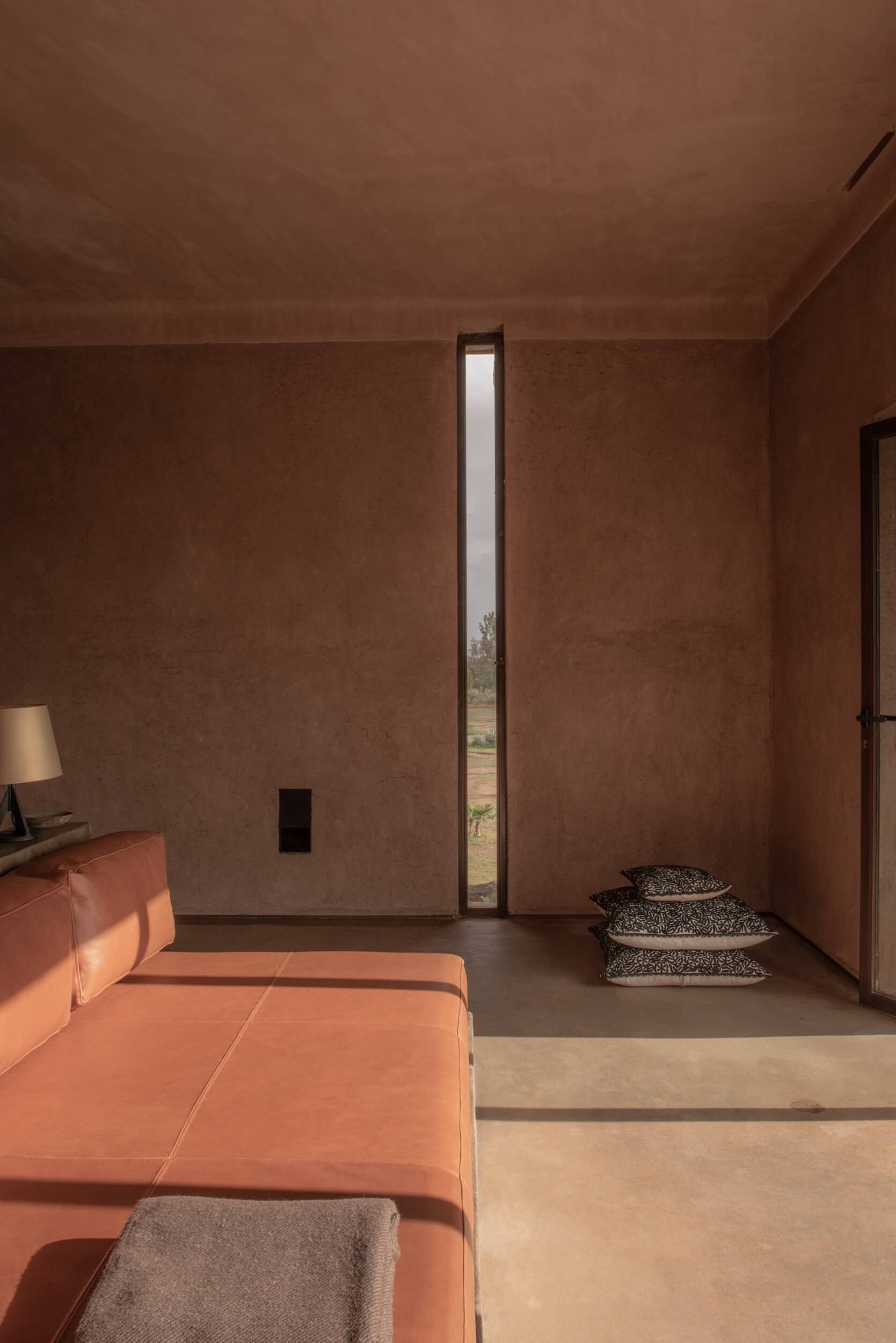 Villa D, Africa — Terracotta-walled bedroom with narrow window framing landscape, coral leather bed, and scattered cushions on elevated platform.