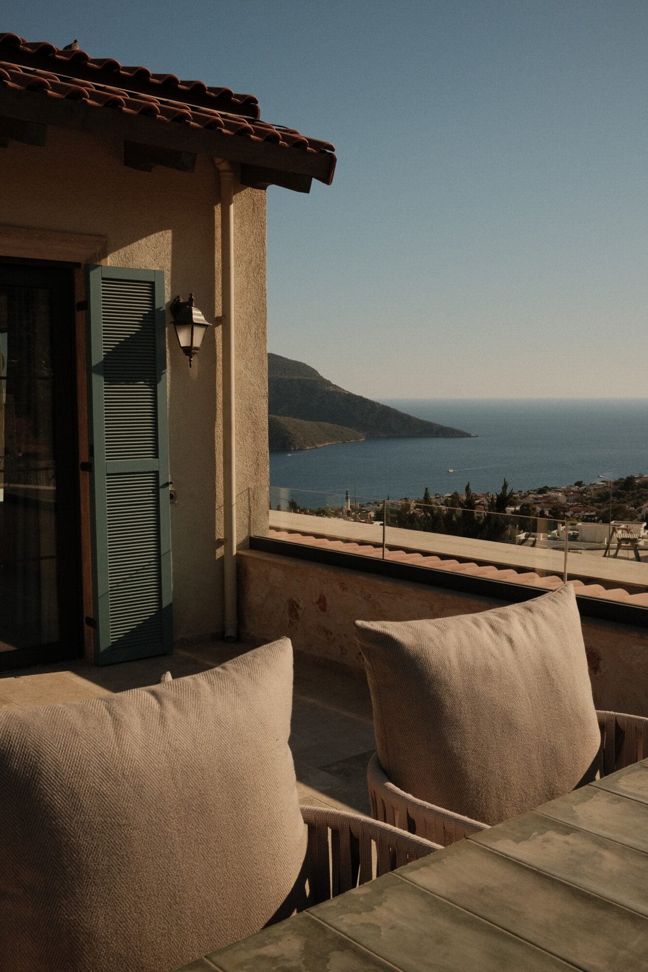 Villa Thalia by Muses Kalkan, Kalkan, Turkey — Terracotta-roofed Mediterranean villa with cream pillows on wooden chairs, green shuttered door, and panoramic sea view overlooking coastal bay at golden hour.