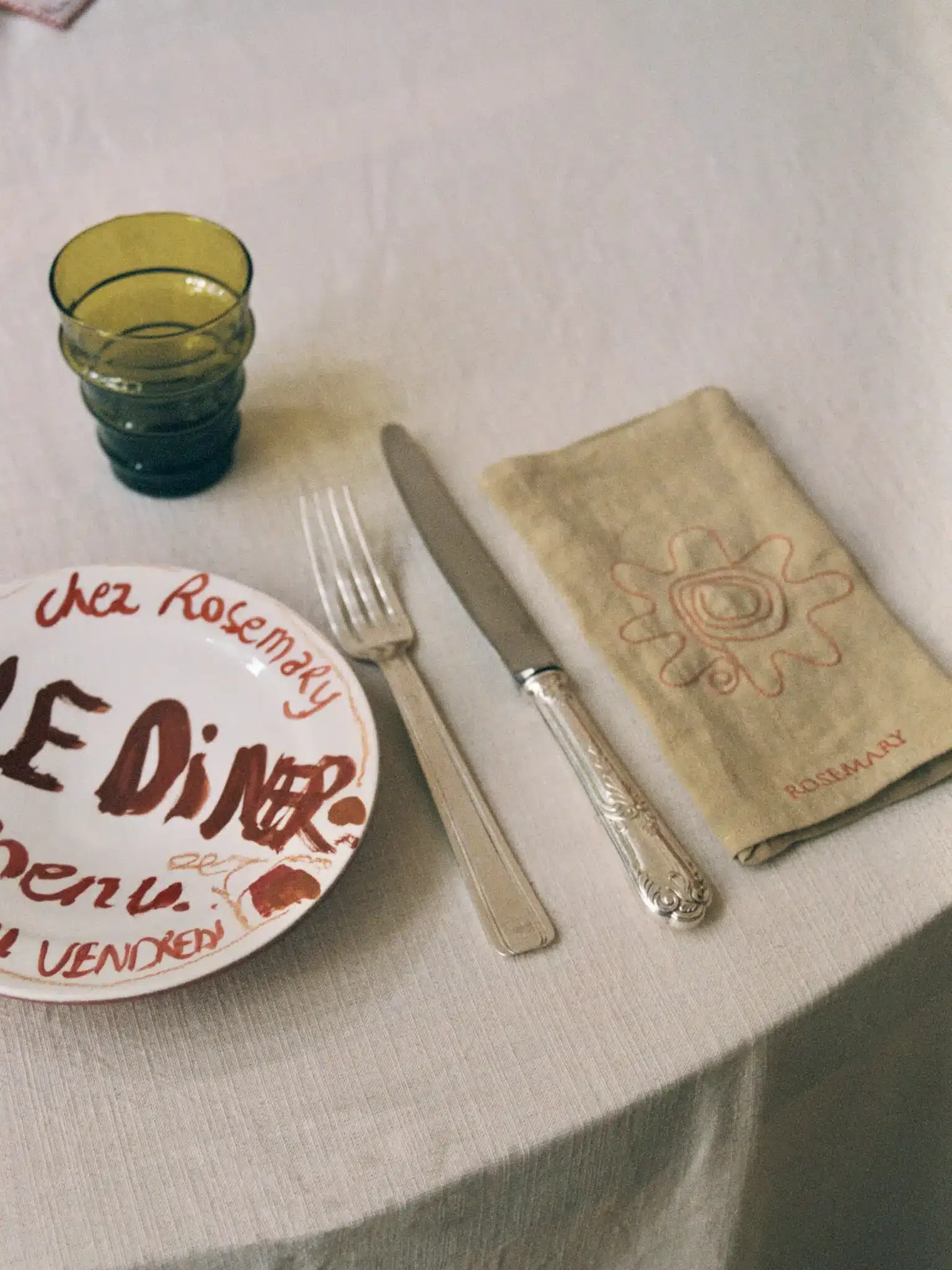 Rosemary, Africa — Elegant place setting with embroidered napkin, silver cutlery, and hand-painted menu on cream tablecloth, soft natural light.