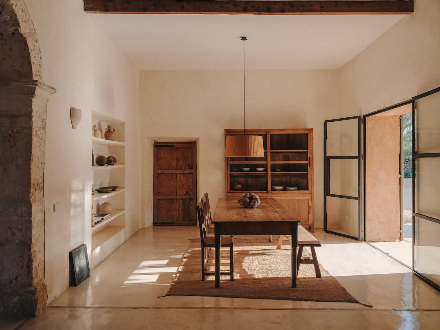 Can Lluïssó, Balearic Islands, Spain — Minimalist dining room with wooden table, terracotta storage cabinets, built-in shelving, and natural light through gridded windows.
