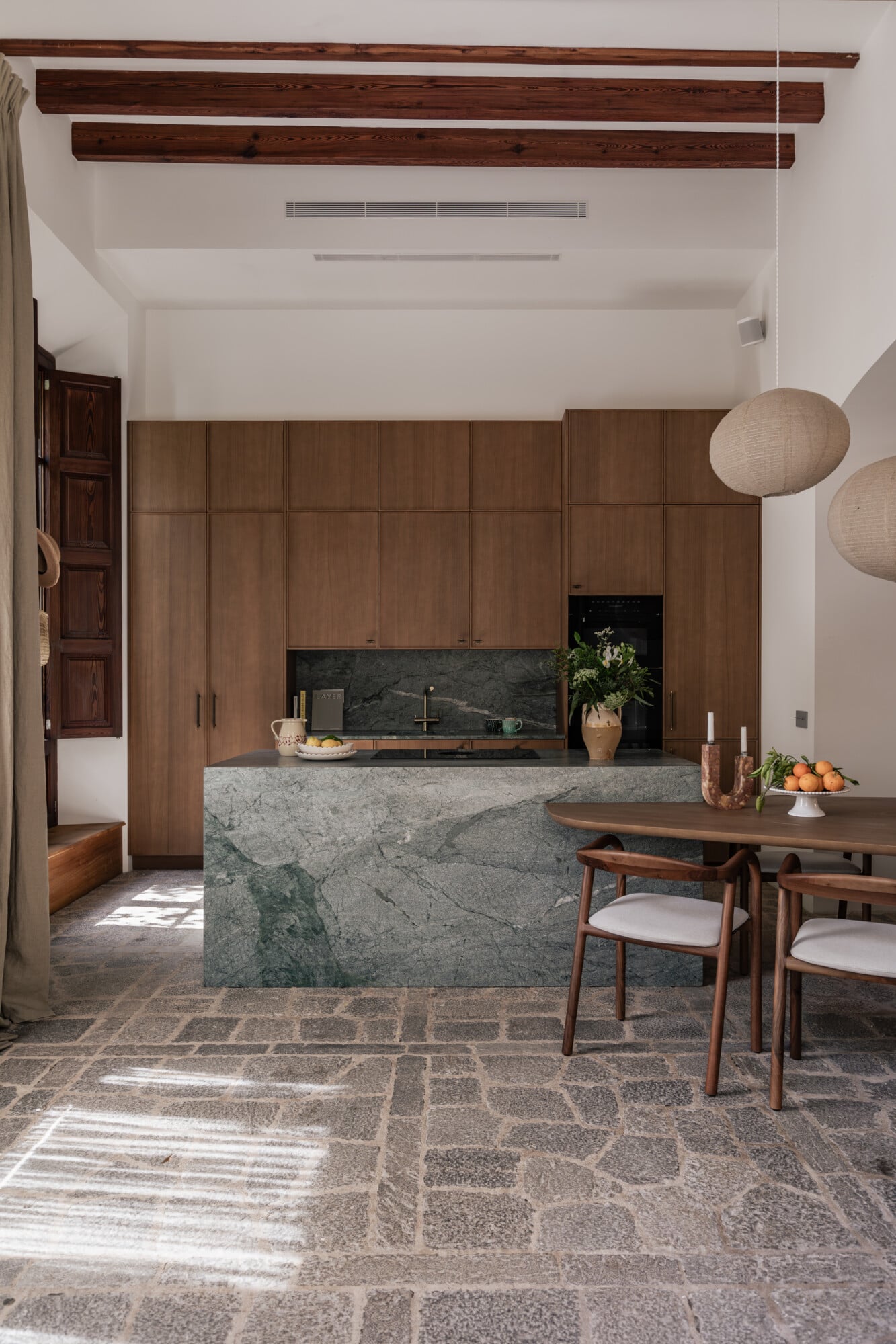 Mon Cor, Balearic Islands, Spain — Walnut cabinetry and green marble kitchen island with dining table, exposed timber beams, natural light, minimalist hospitality interior.