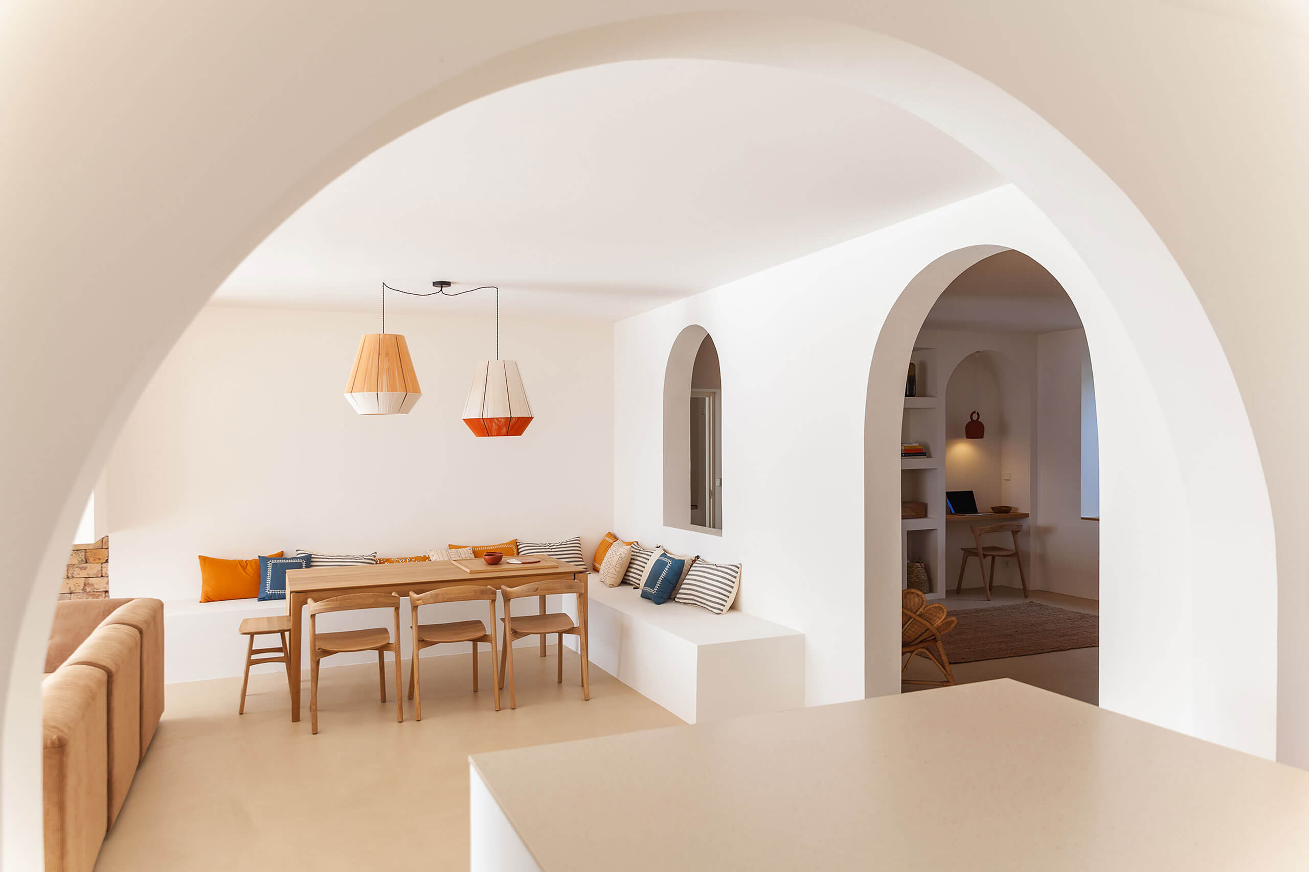 Casa Santa Teresa, Europe — Whitewashed dining room with curved archways, wooden furniture, orange pendant lights, and built-in bench seating with blue and orange pillows.