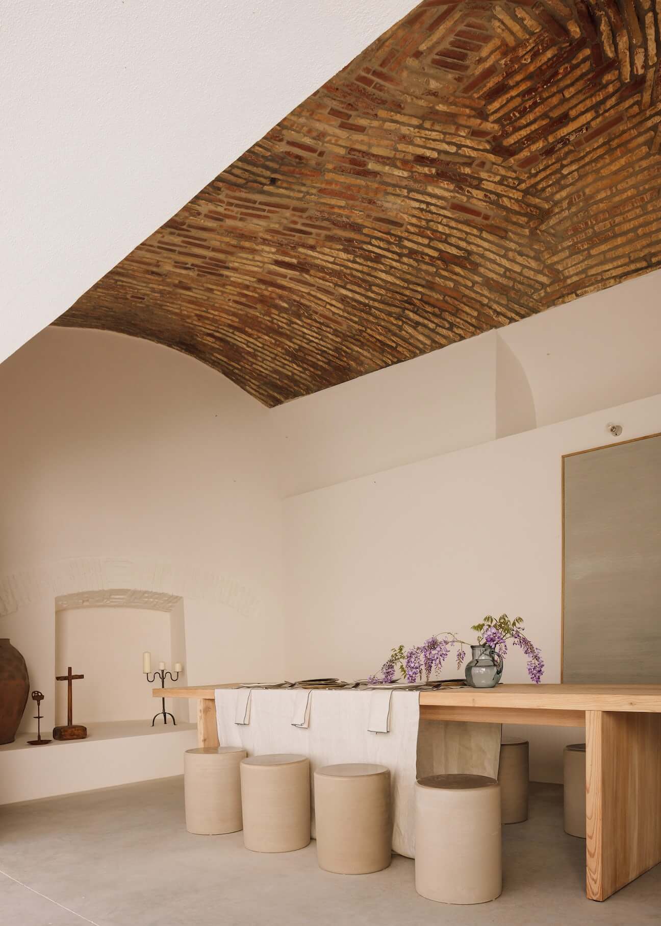 Casa Quatro, Europe — Minimalist bathroom with exposed wooden ceiling beams, cream cylindrical seating, natural light, and wisteria in ceramic vessel.