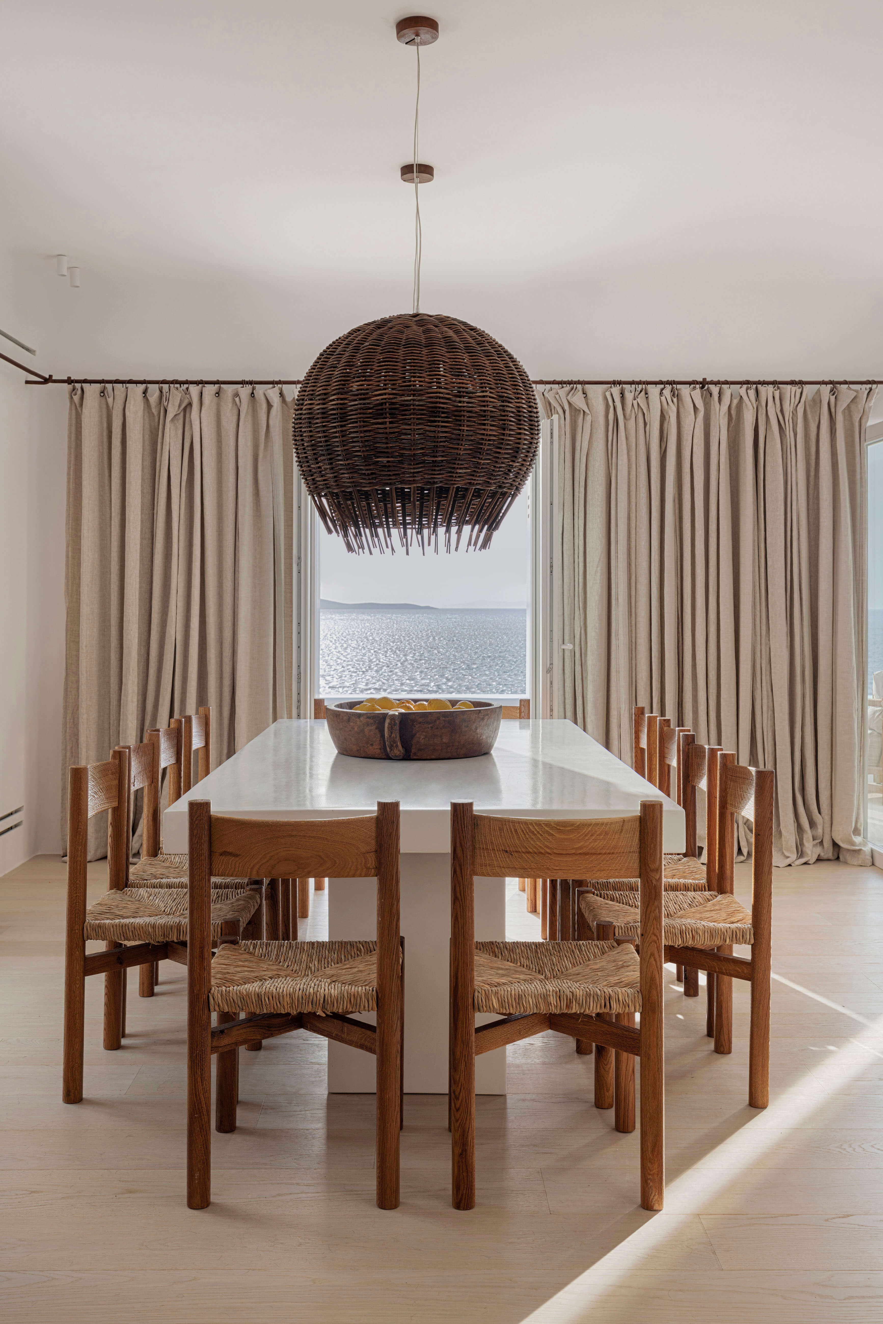 Villa Tama, Europe — Dining room with woven pendant light, wooden chairs, white table, and sea view through curtained windows.