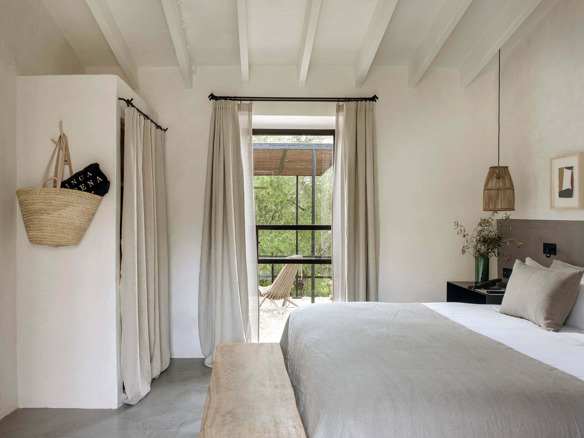 Finca Serena, Balearic Islands, Spain — Minimalist bedroom with exposed beam ceiling, linen curtains, pendant lighting, and glass doors opening to terrace with sea views.