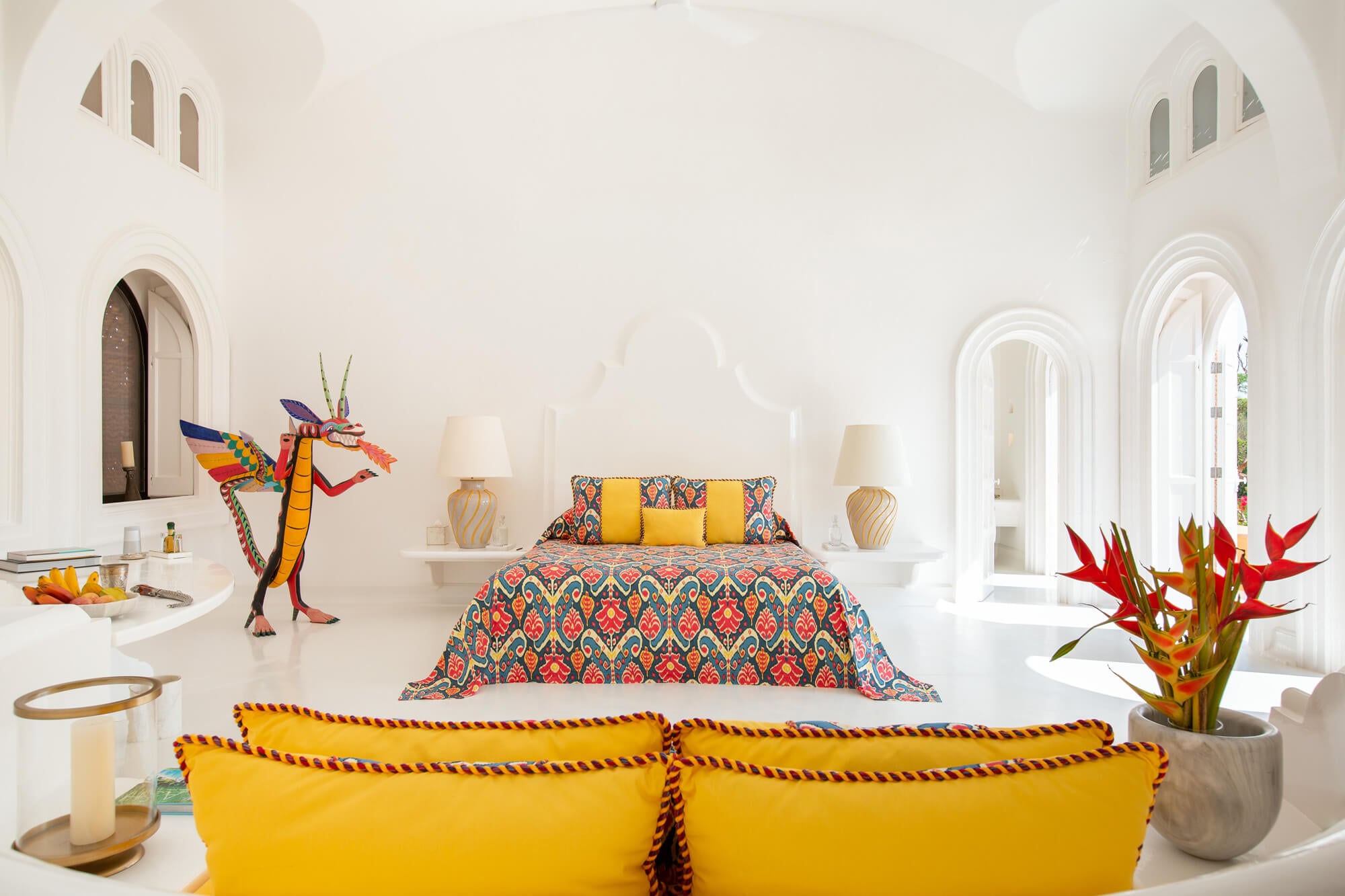 Cuixmala, Mexico — Bright bedroom with arched windows, colorful patterned bedspread, yellow cushions, woven dragon sculpture, and tropical flowers in minimalist white interior.