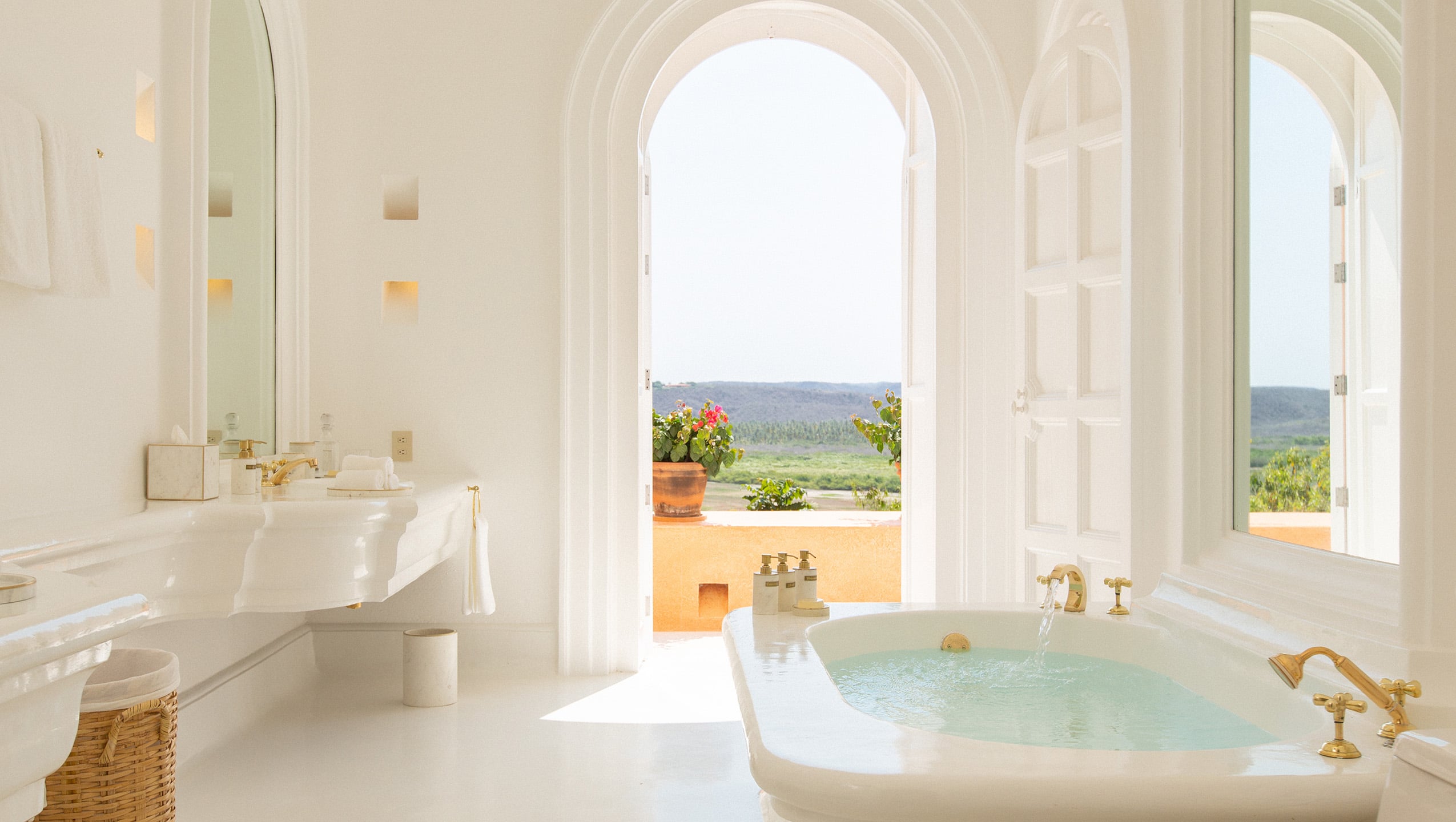 Cuixmala, Mexico — Minimalist bathroom with arched doorway, soaking tub, brass fixtures, overlooking forested coastline and terracotta terrace.