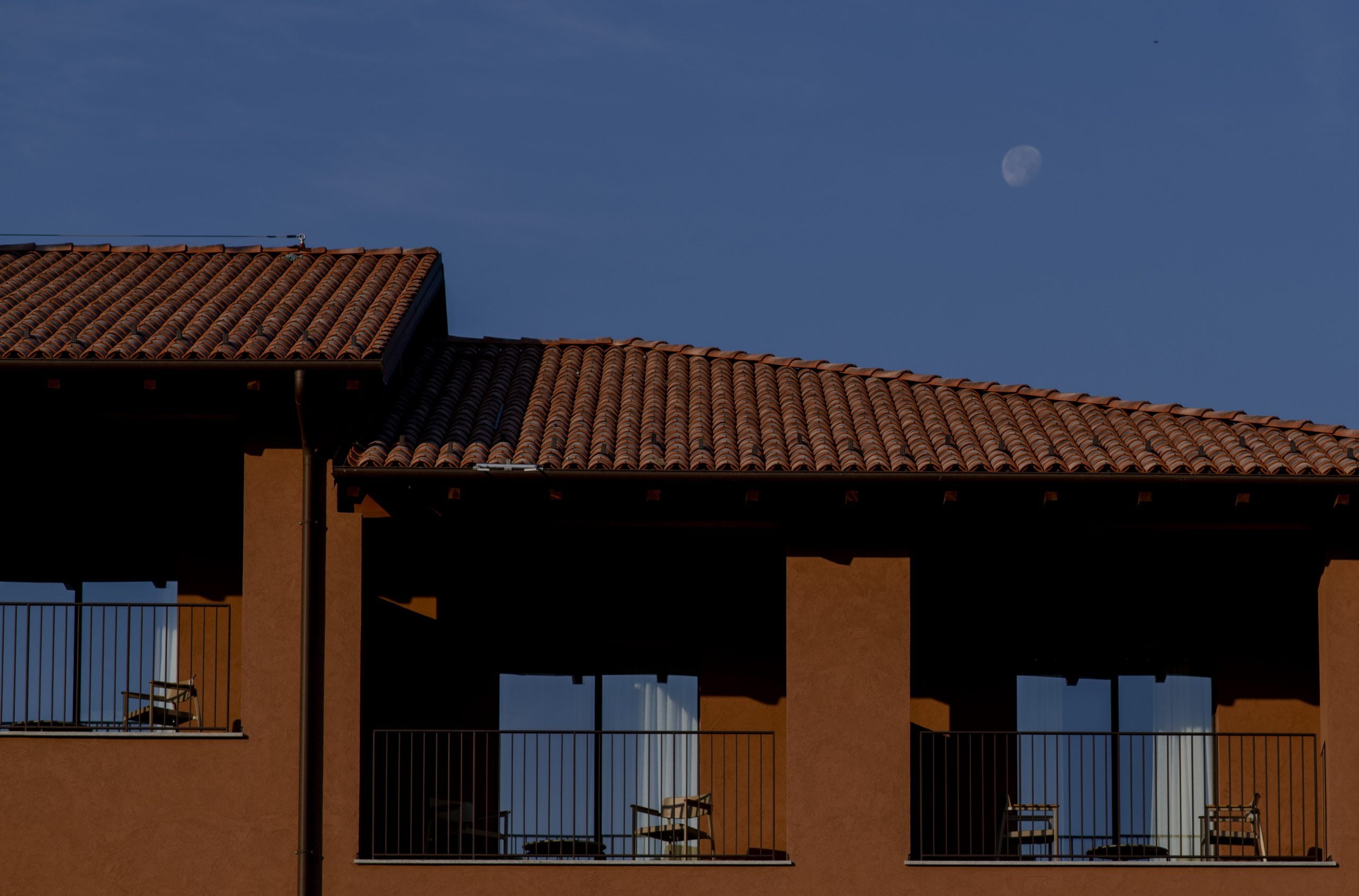 Casa di Langa, Europe — Terracotta-roofed building with ochre stucco facade, steel-railed balconies, dramatic shadows, moonlit sky at dusk.